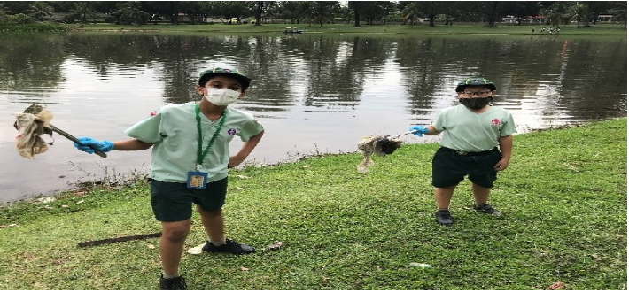 Two children in green uniforms and masks hold trash-picking tools near a pond in a park.