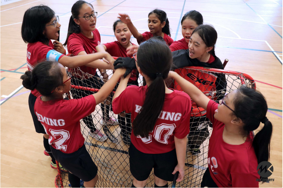 Girls in red jerseys celebrate in a team huddle around a hockey net on an indoor gym floor.