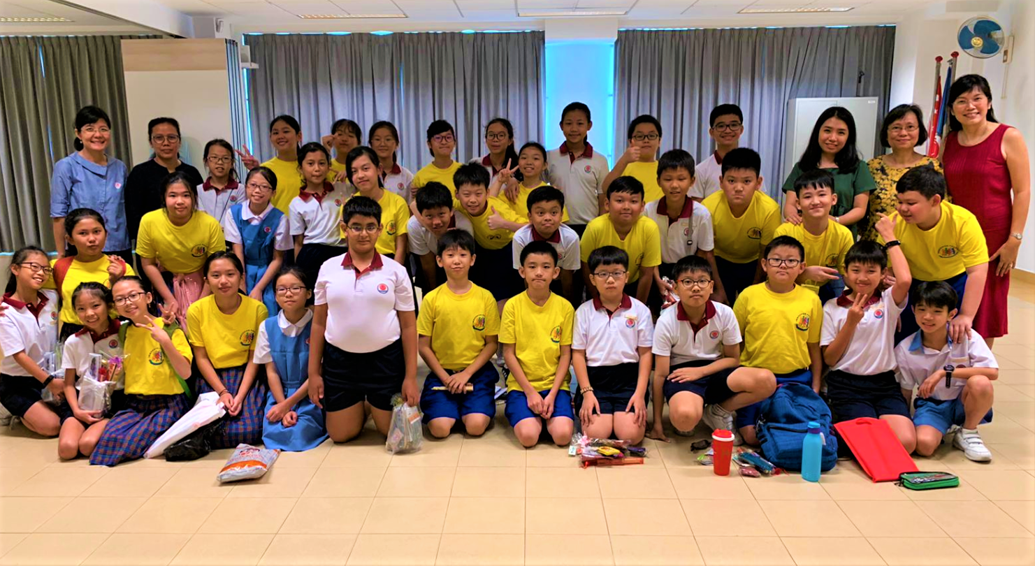 A large group of school children in yellow and white uniforms pose with several adult teachers indoors.