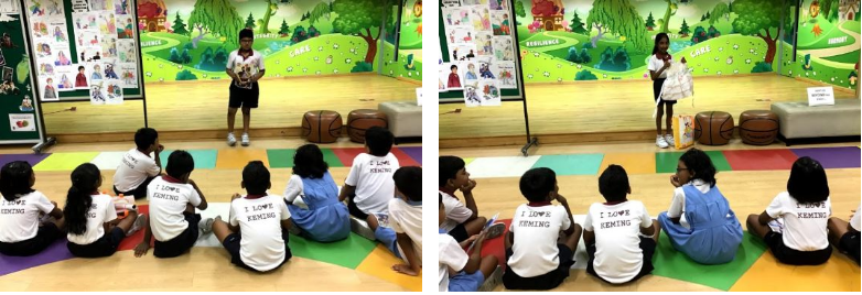 Children in matching shirts sit on a colorful mat facing a student presenting in a decorated classroom.