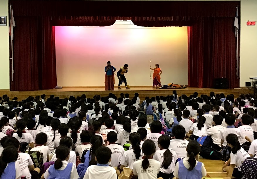 Three performers in colorful costumes act on a stage before a large seated audience of children in white shirts.
