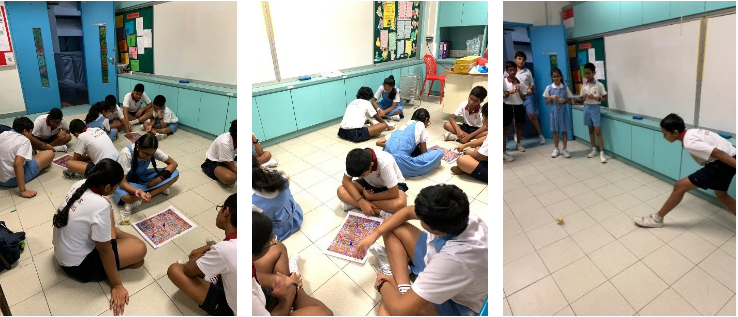 Students in white uniforms sit on a classroom floor examining large colorful printed sheets, while others stand nearby.