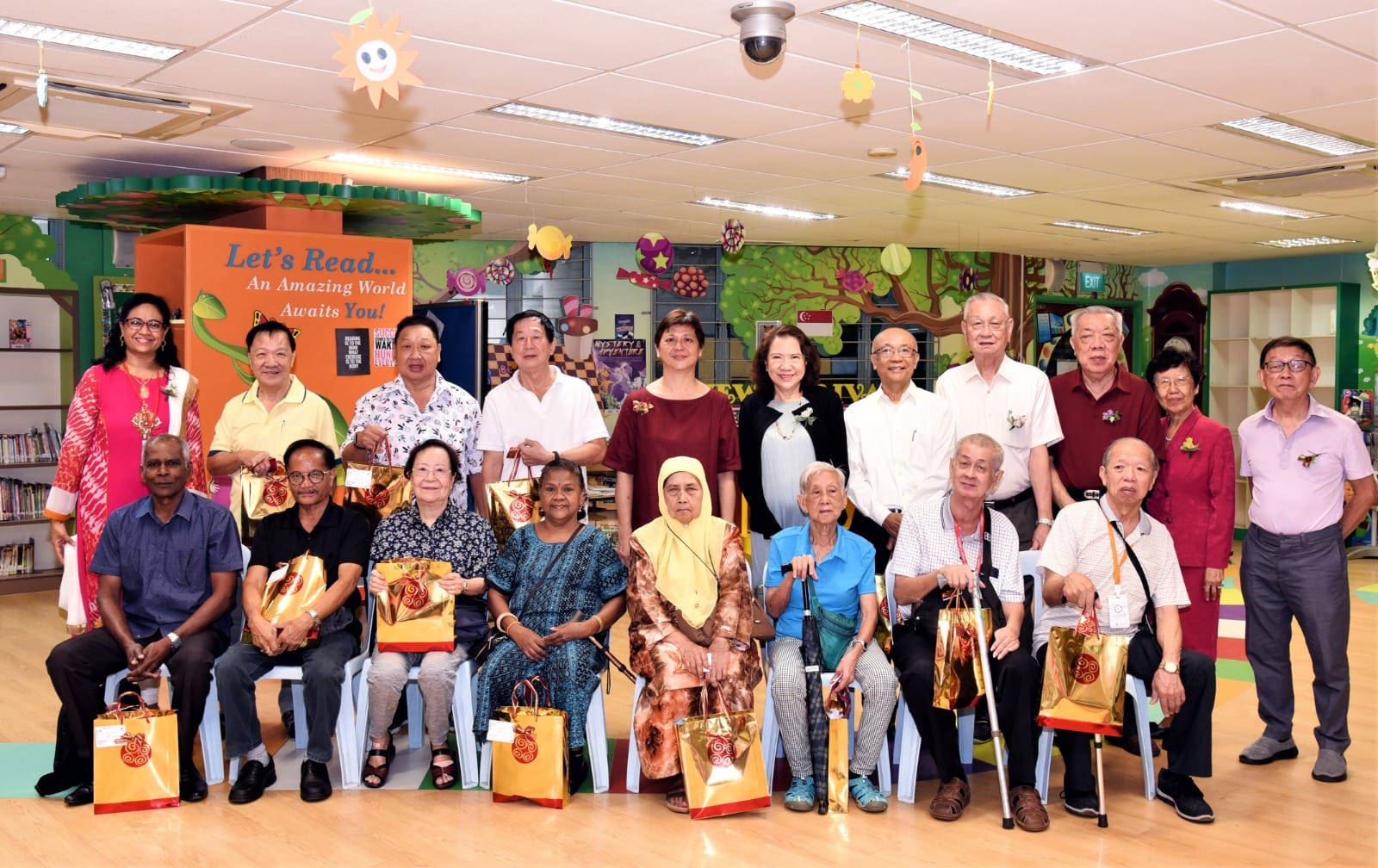 A large group of elderly and middle-aged adults pose together in a colorfully decorated library, holding gold gift bags.