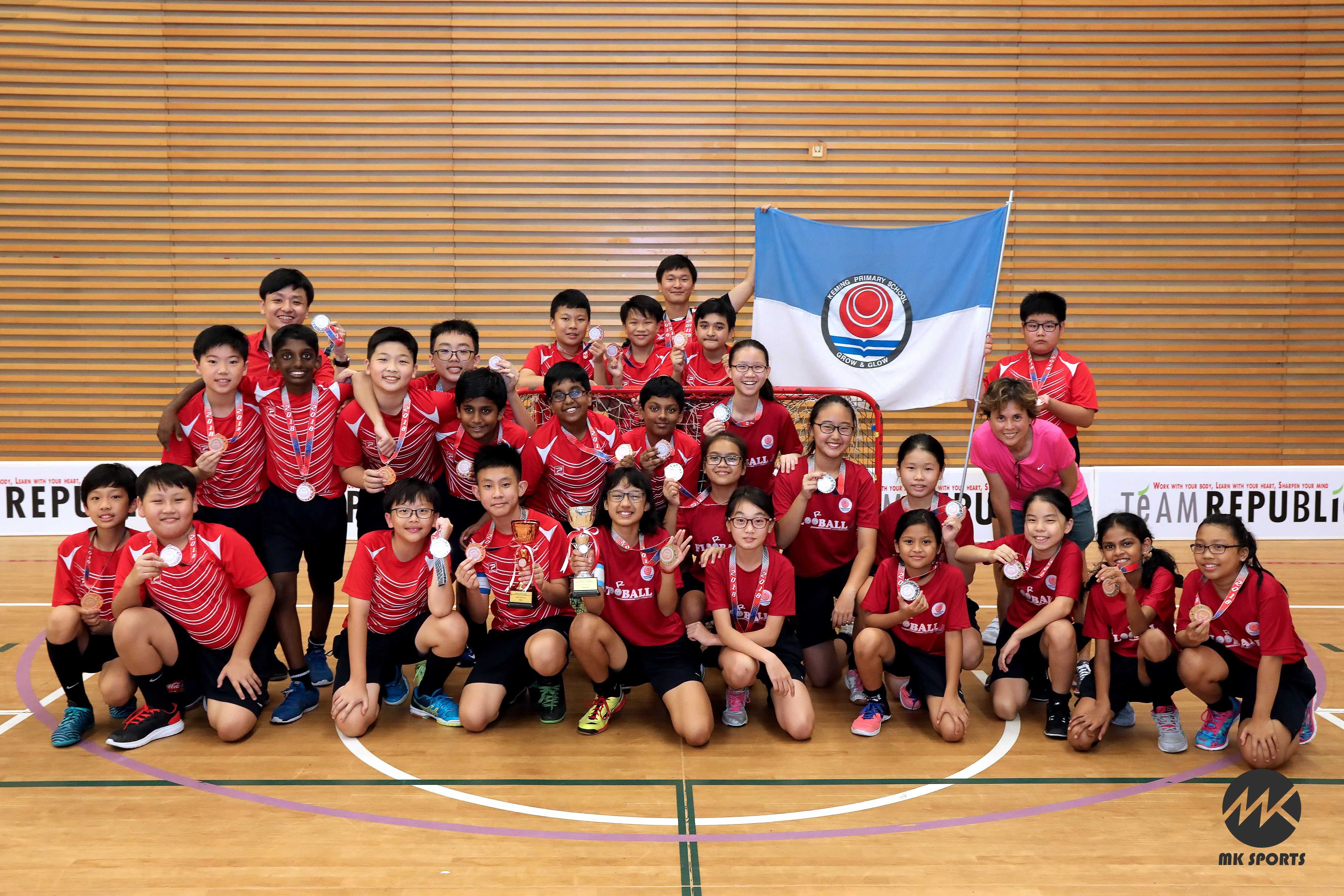 Group of children in red sports uniforms posing with medals, trophies, and a school flag inside an indoor sports hall.