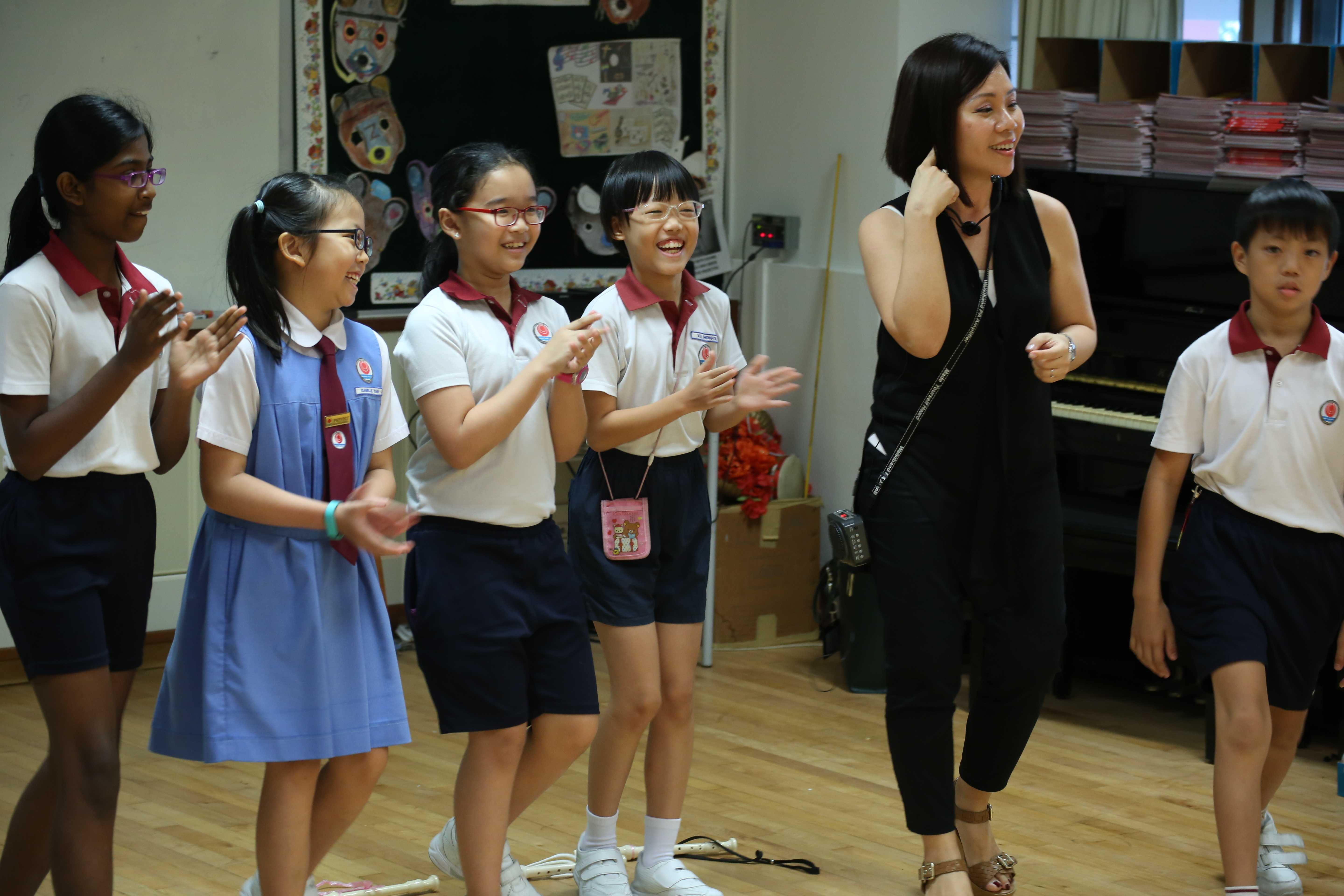 A teacher in black gestures while five uniformed primary school children clap and smile in a music classroom with a piano.