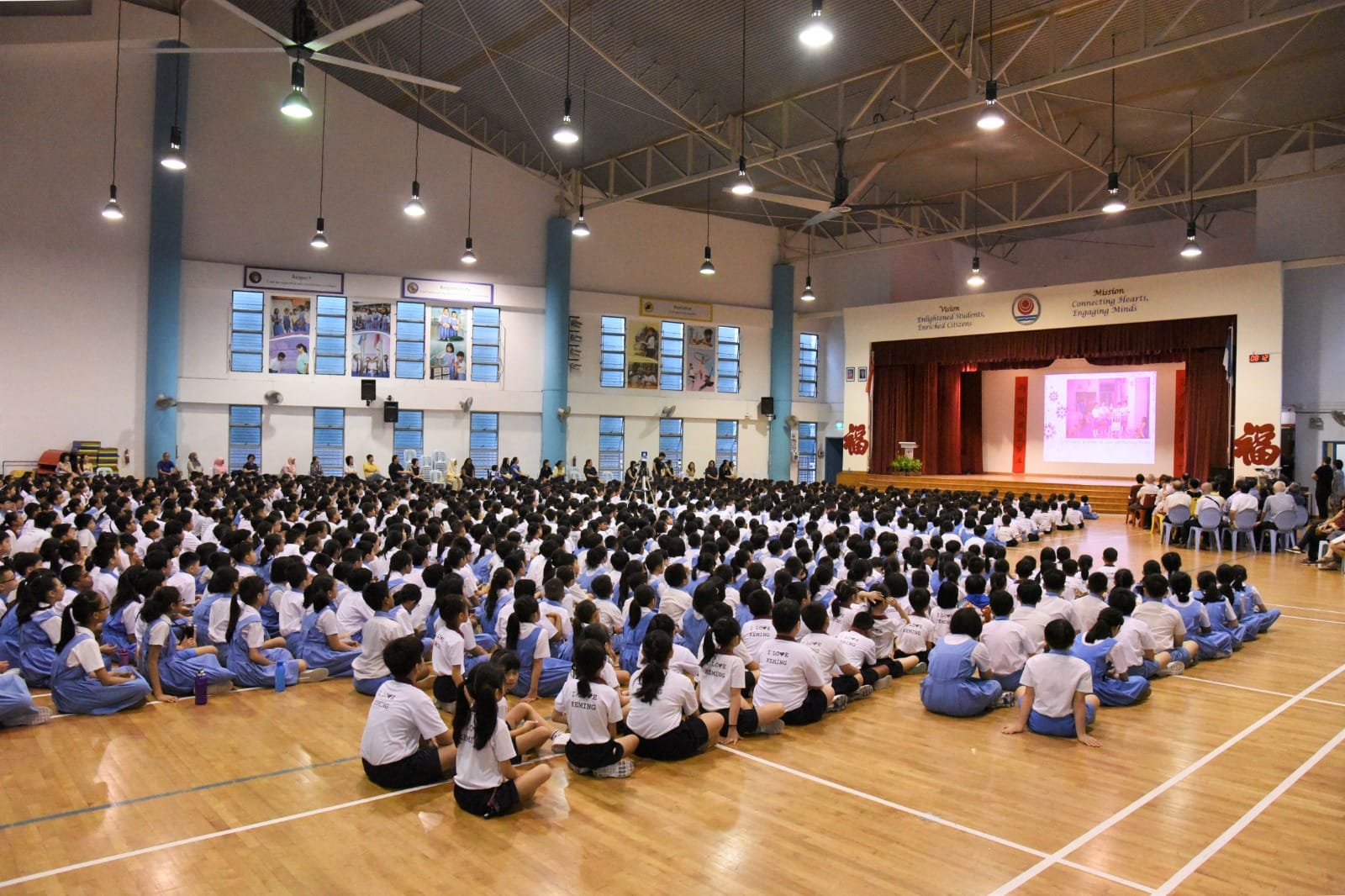 Hundreds of students in white and blue uniforms sit on a school hall floor facing a lit stage with a projected screen.