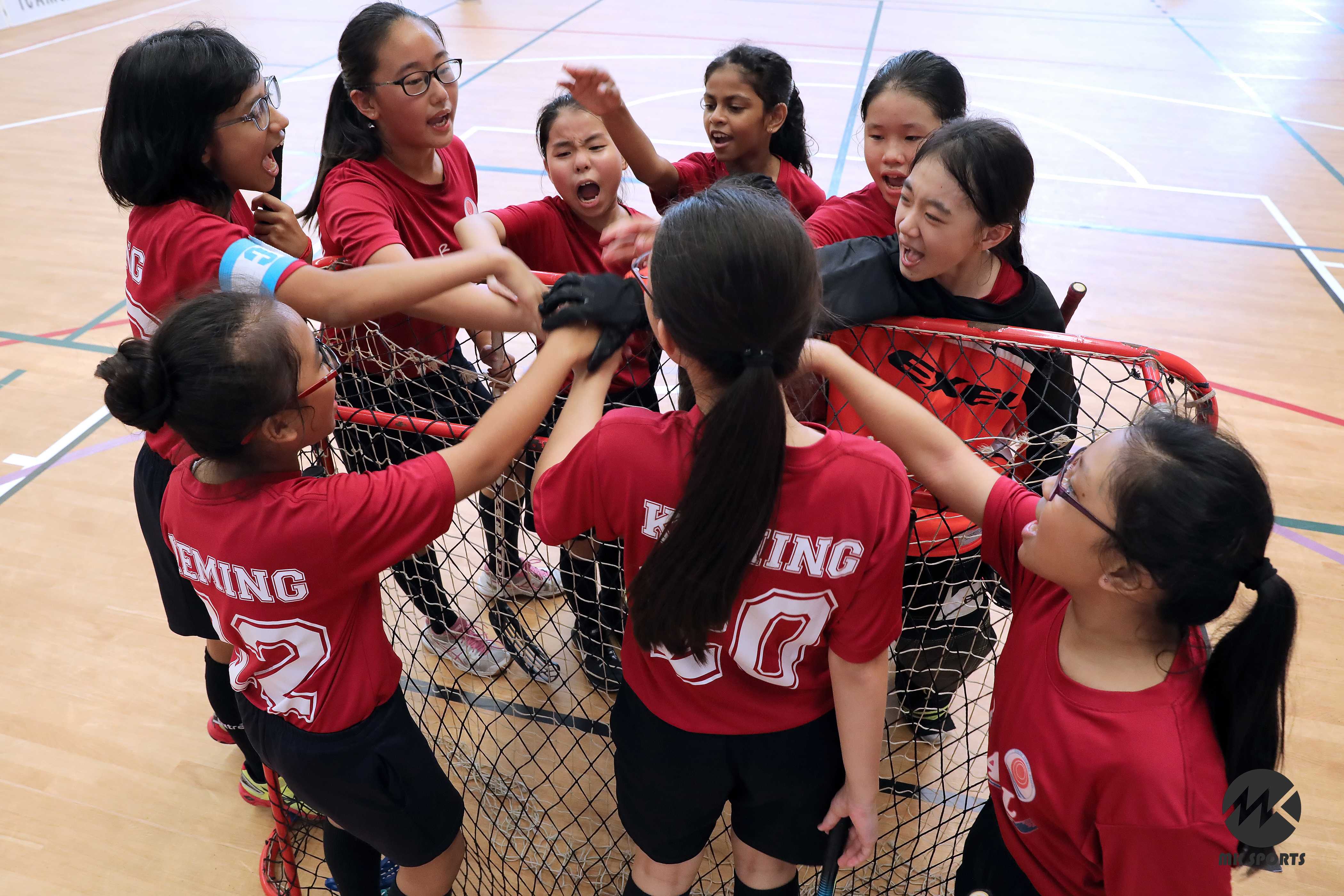 A group of girls in red jerseys celebrate together around a floorball goal on an indoor sports court.