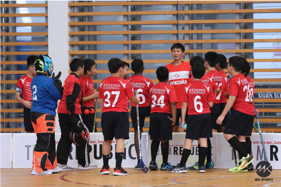 Youth floorball team in red Keming Floorball jerseys huddles with a coach on an indoor court.