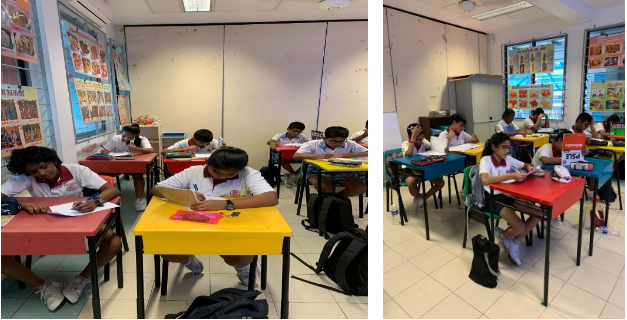Students in white uniforms sit at colorful desks writing in a bright classroom with posters on the walls.