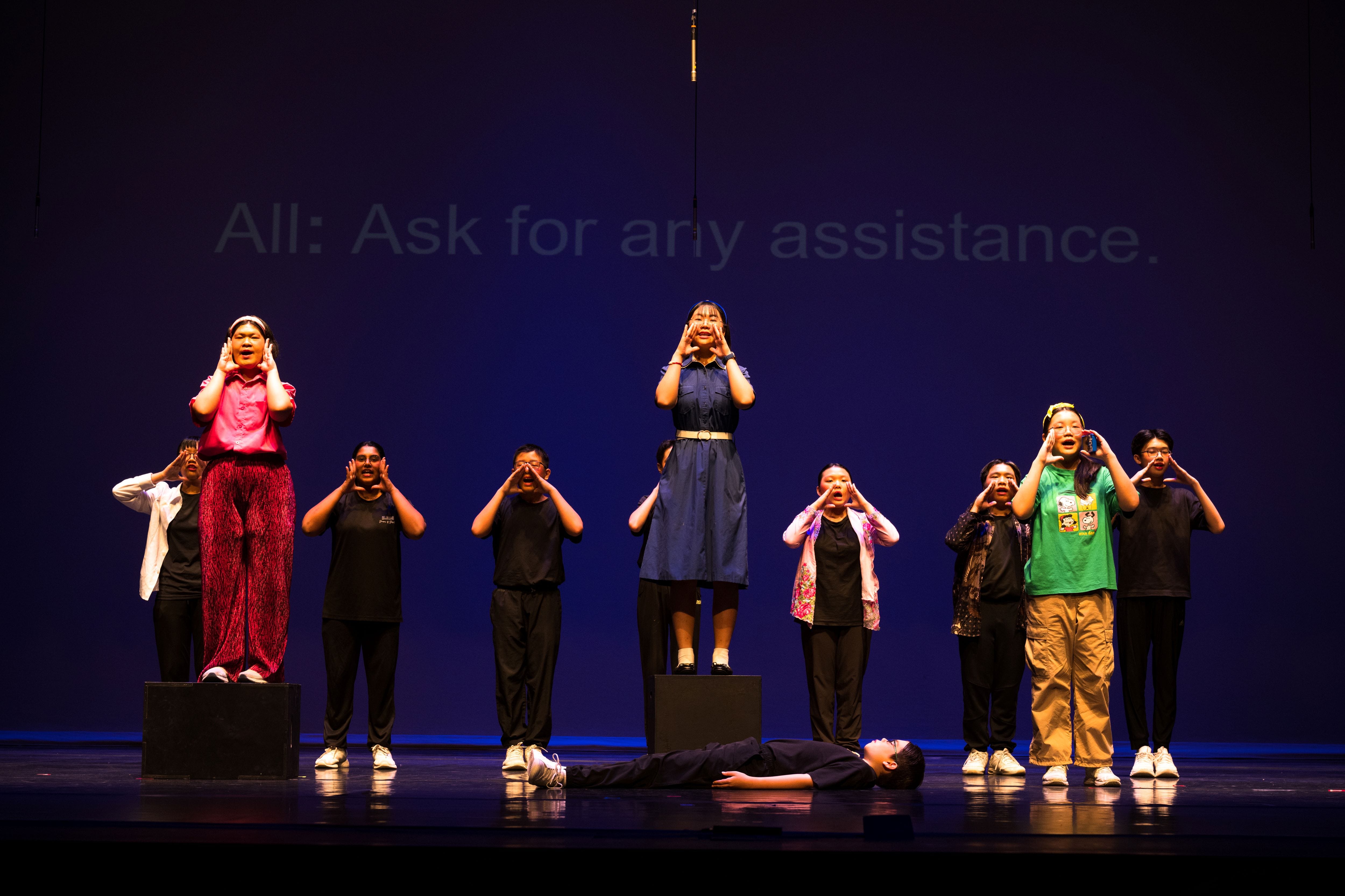 Group of young performers on a dark stage cupping hands to mouths, with one person lying on the floor below.