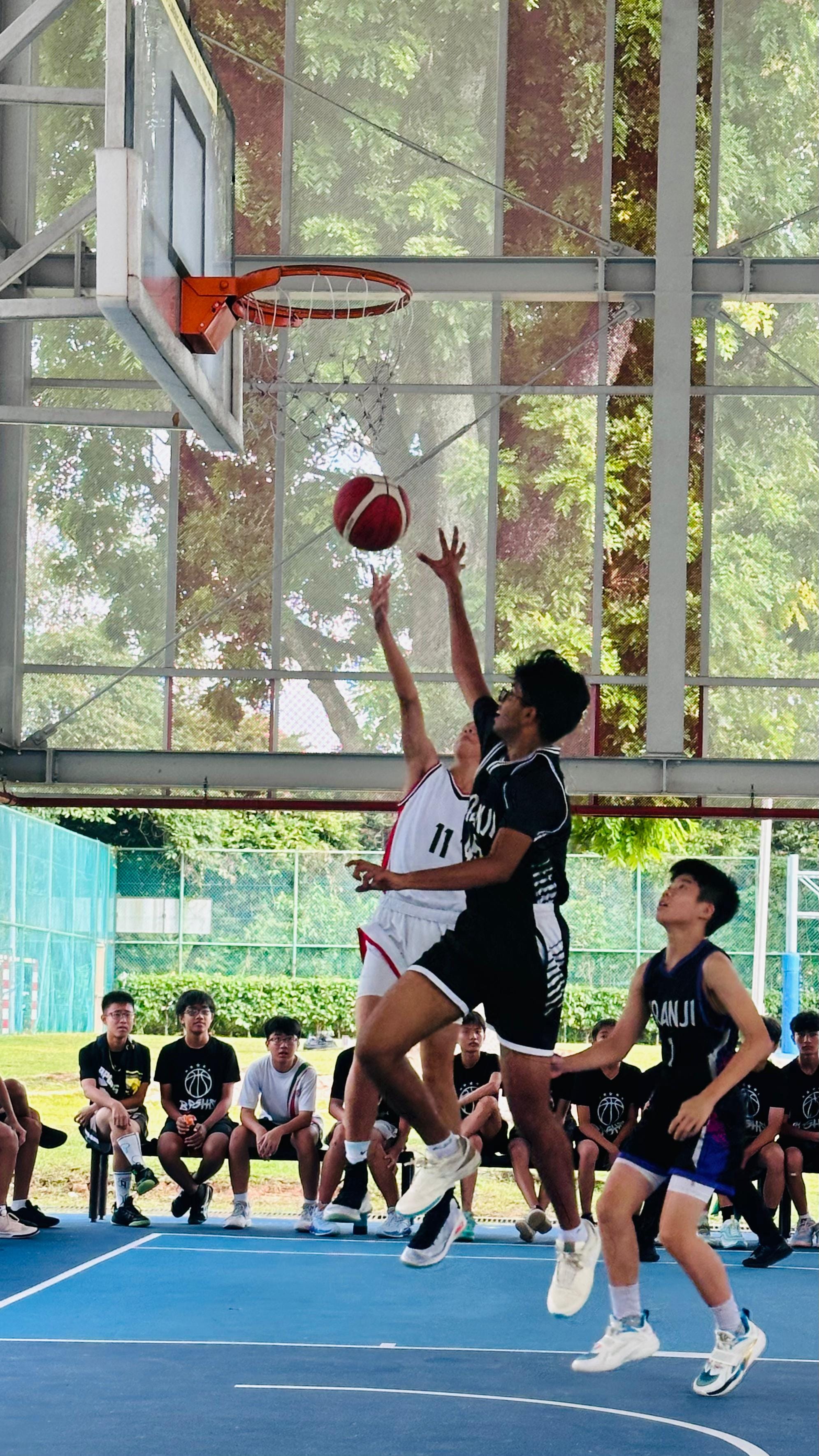 Two players leap toward a basketball hoop during an outdoor game as teammates watch from the sideline.
