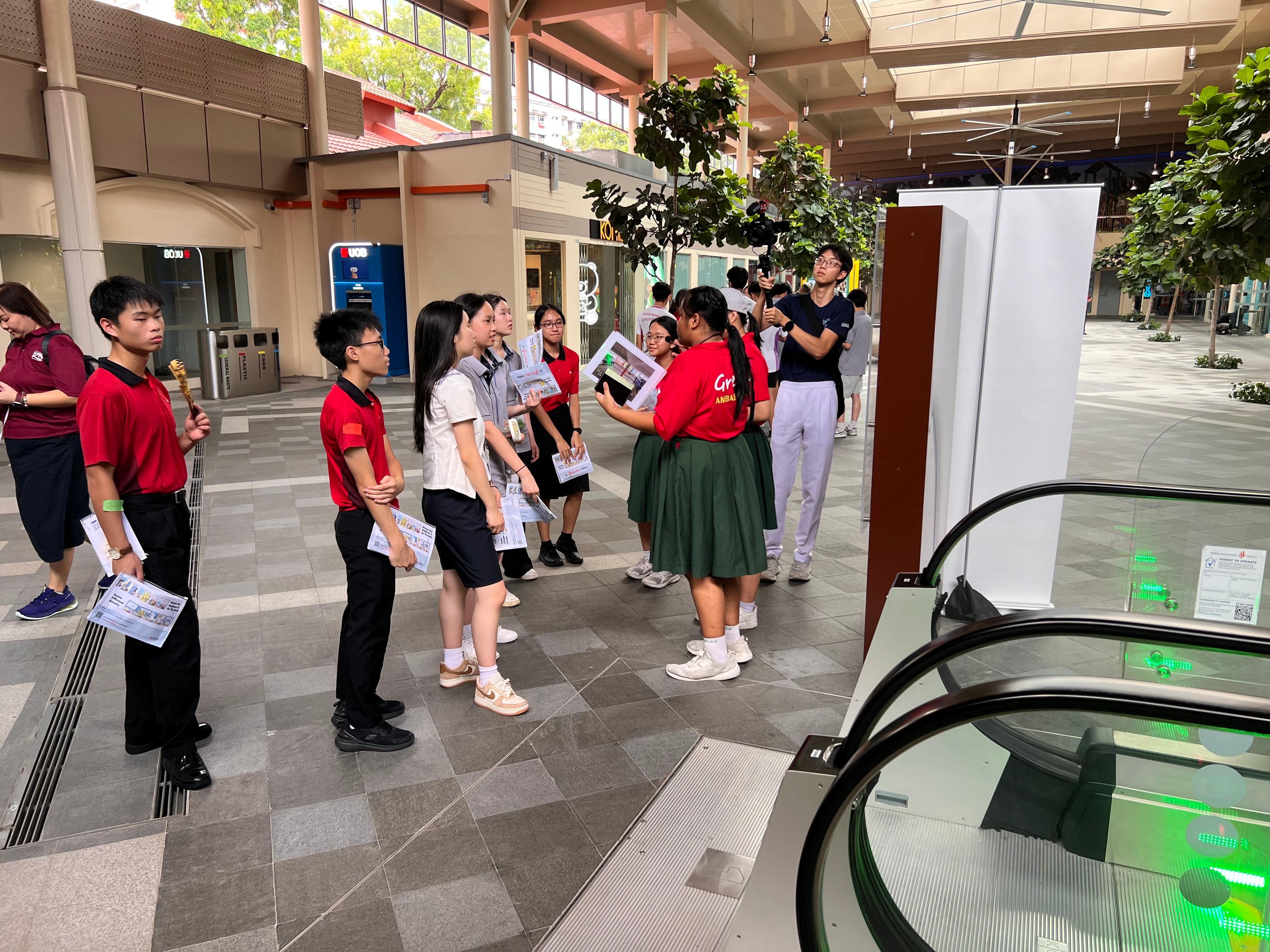Students in red uniforms gather in an indoor public space near an escalator, holding printed materials.