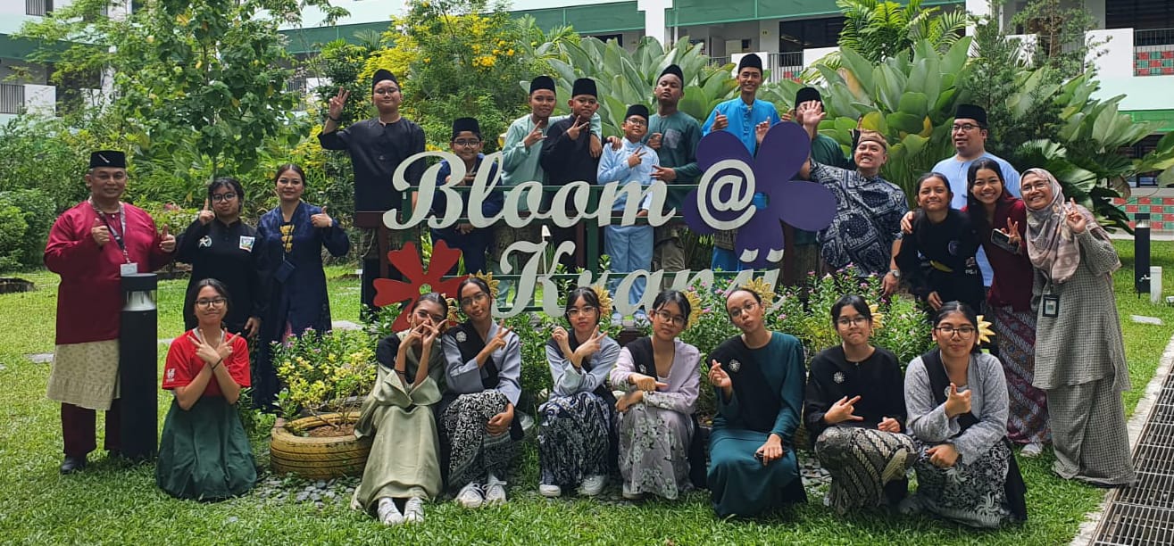 A large group of students and teachers in traditional attire posing outdoors near a sign reading Bloom @ Kranji.