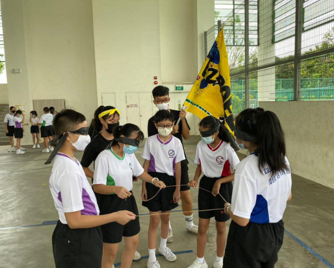 Students in masks and blindfolds hold a rope together indoors during a team-building activity, with a yellow flag nearby.