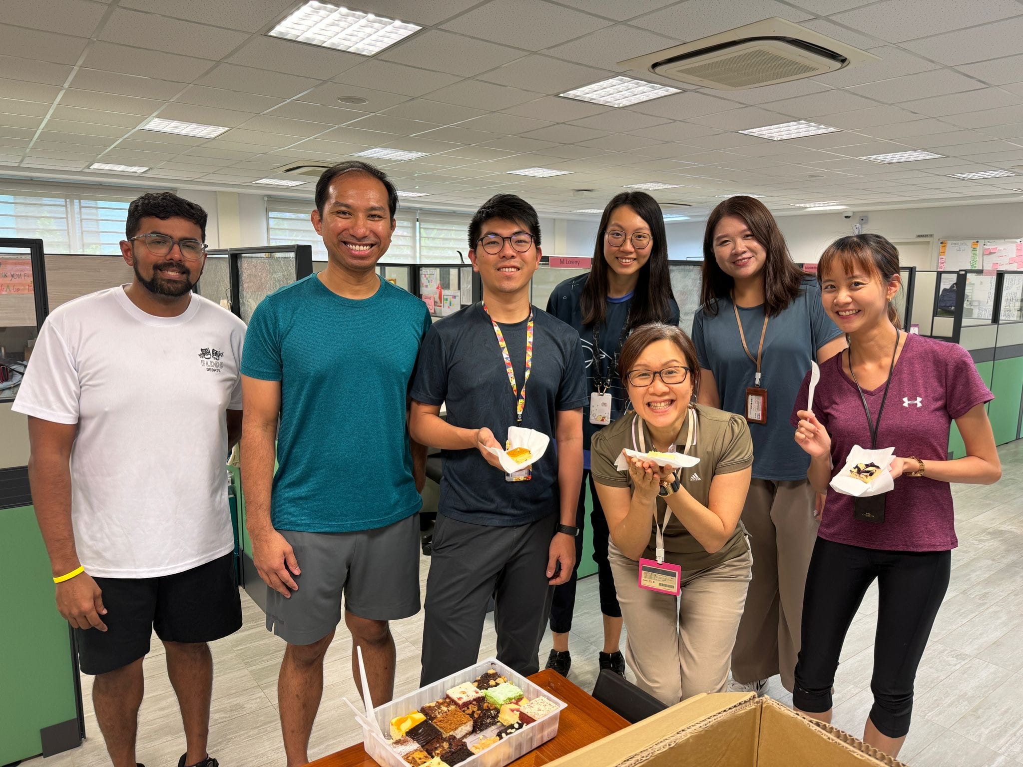 Seven smiling coworkers gather around a tray of assorted cakes in an office, some holding slices on paper plates.
