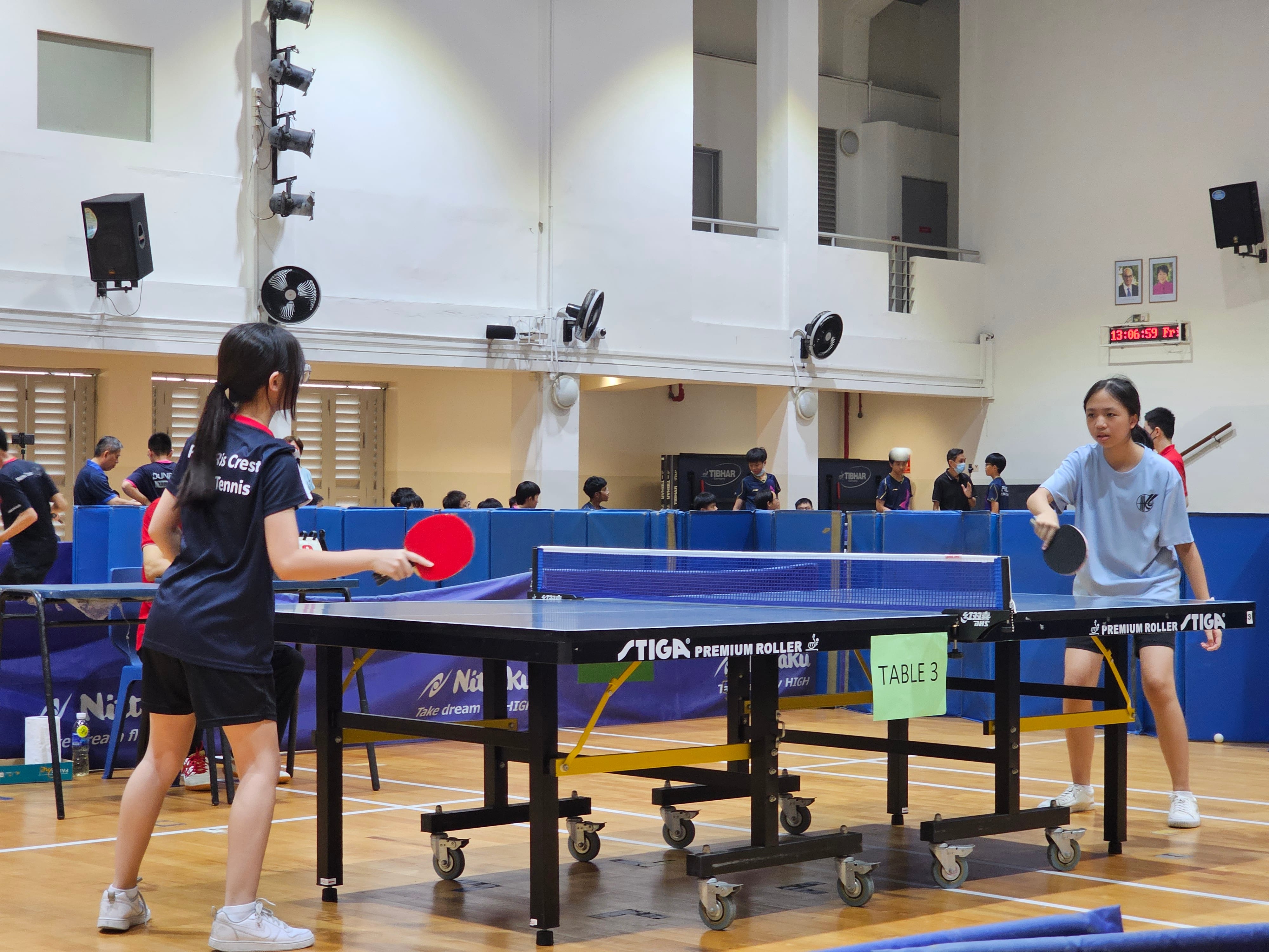 Two young women play table tennis on a Stiga table labeled Table 3 in an indoor sports hall.