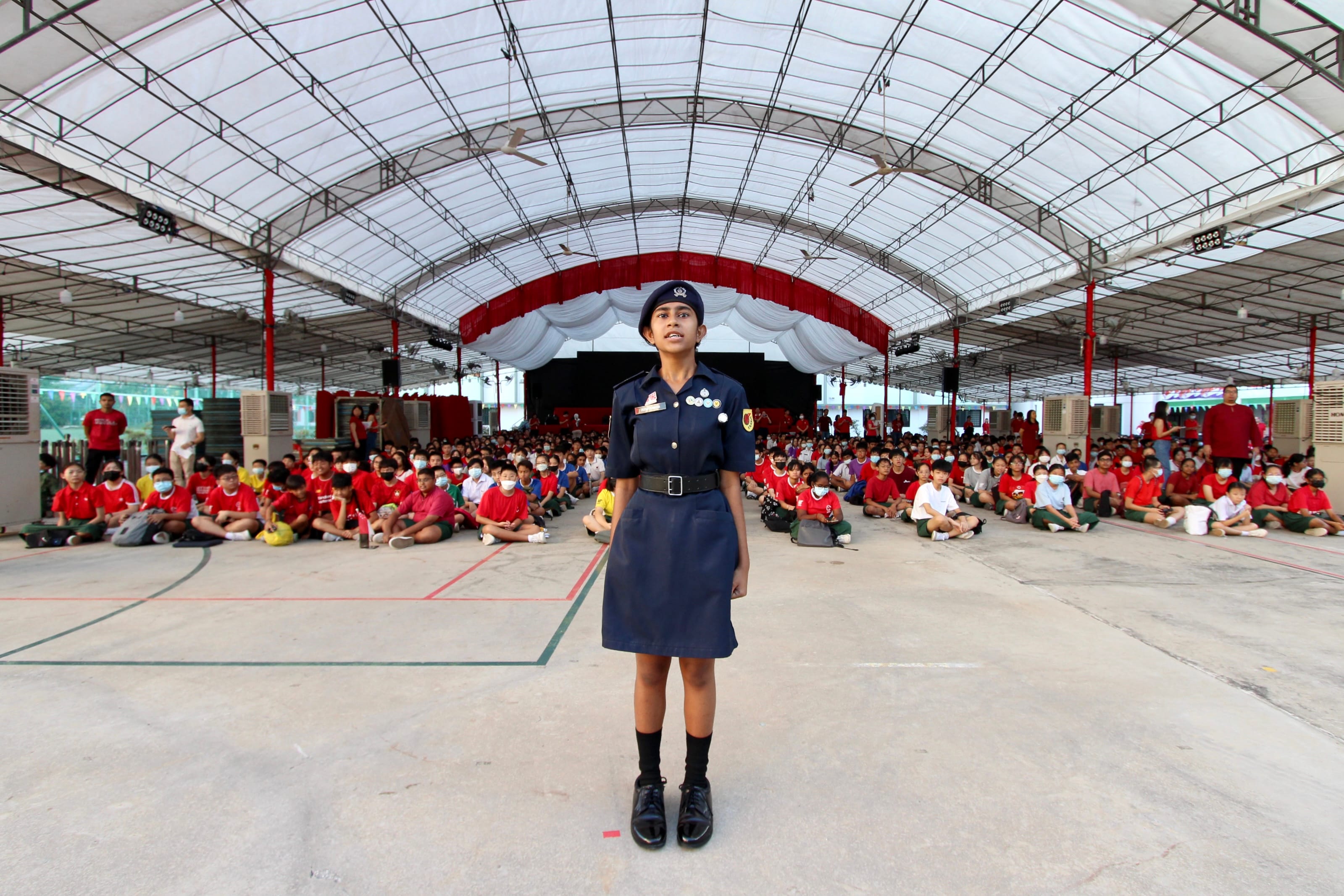 A girl in a navy uniform and beret stands at attention before a large seated crowd of students in a covered hall.