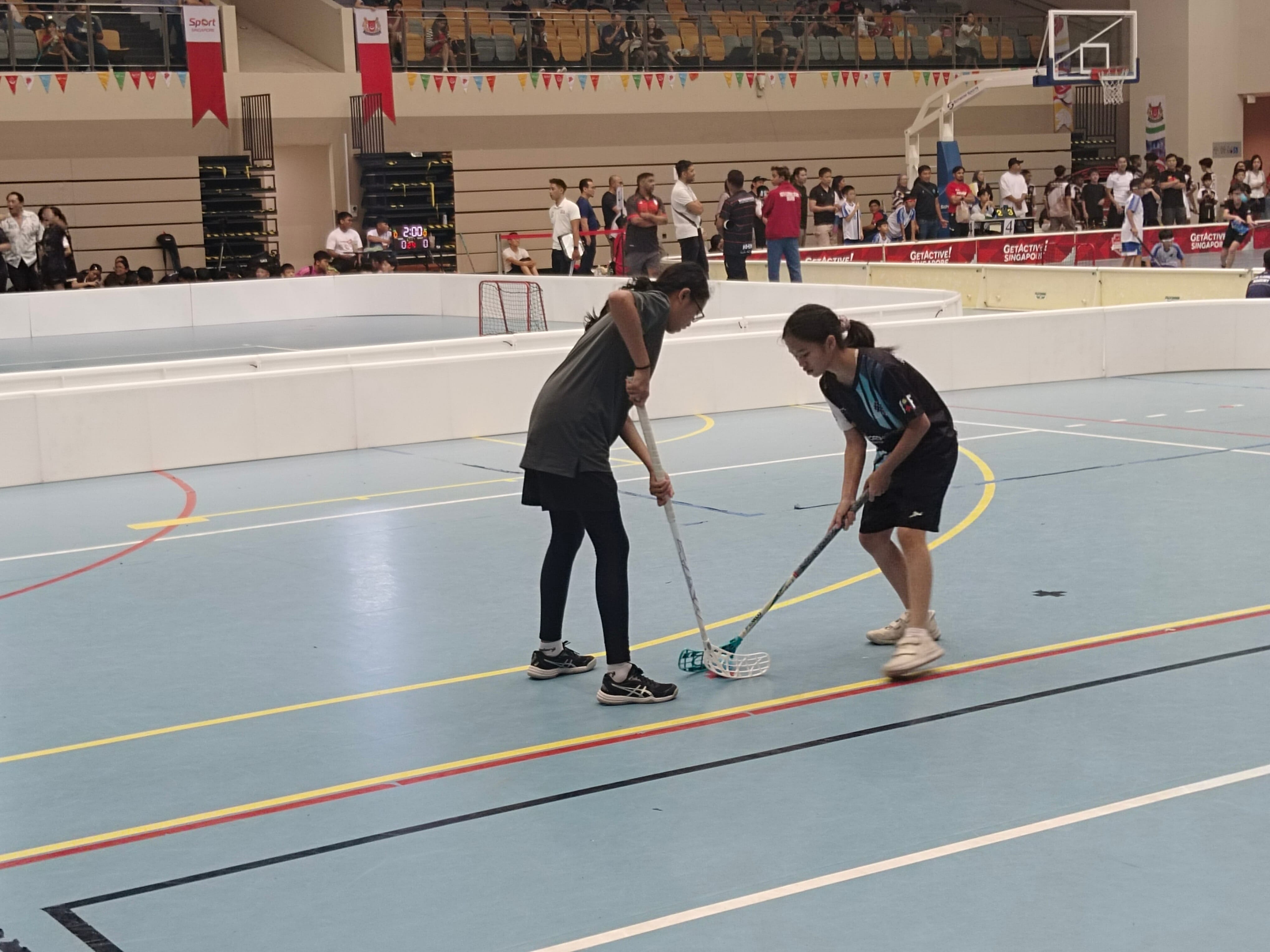 Two young women compete in floorball inside a sports hall, leaning over sticks near the ball.