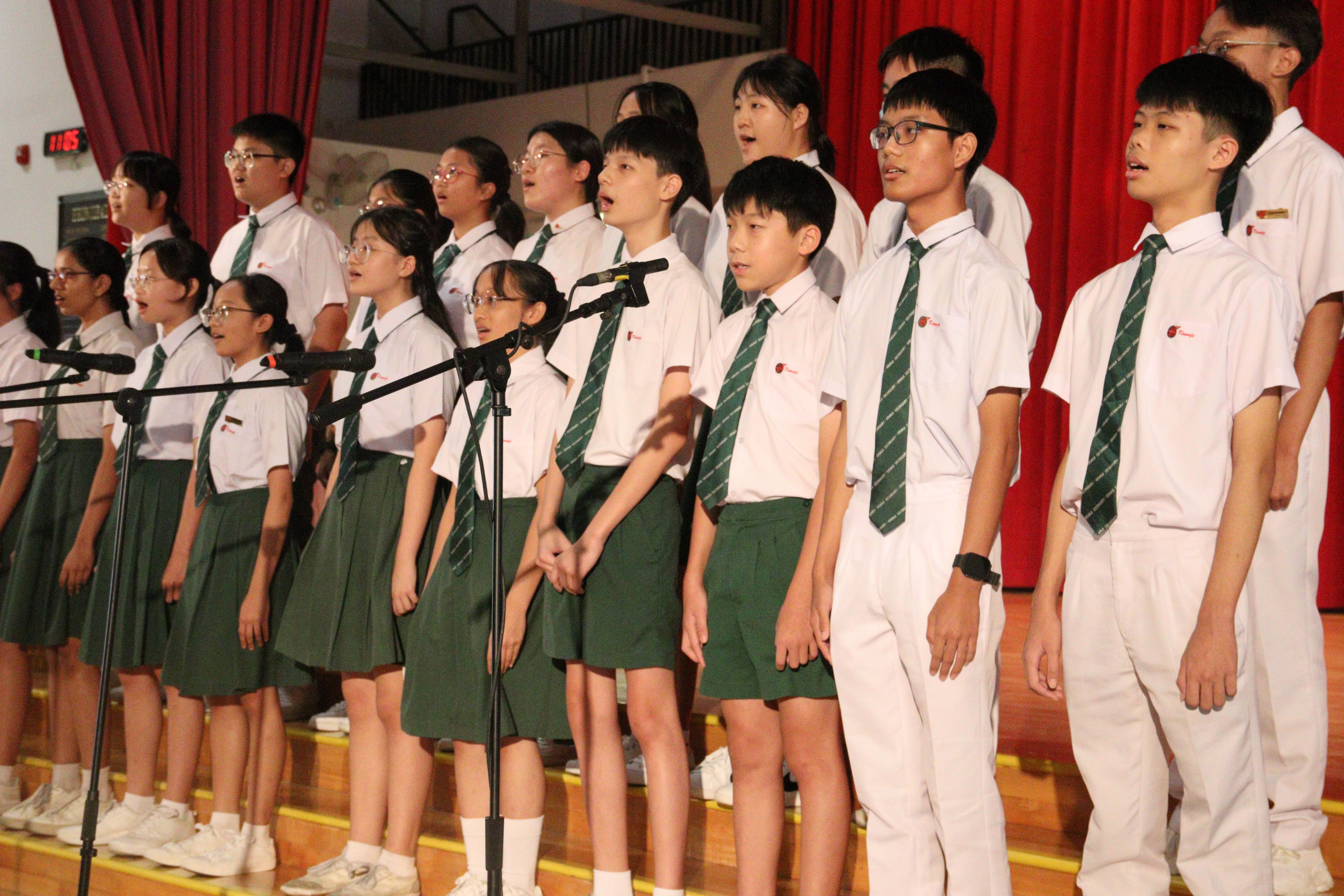 Students in white shirts and green ties sing in a choir on a stage with red curtains behind them.