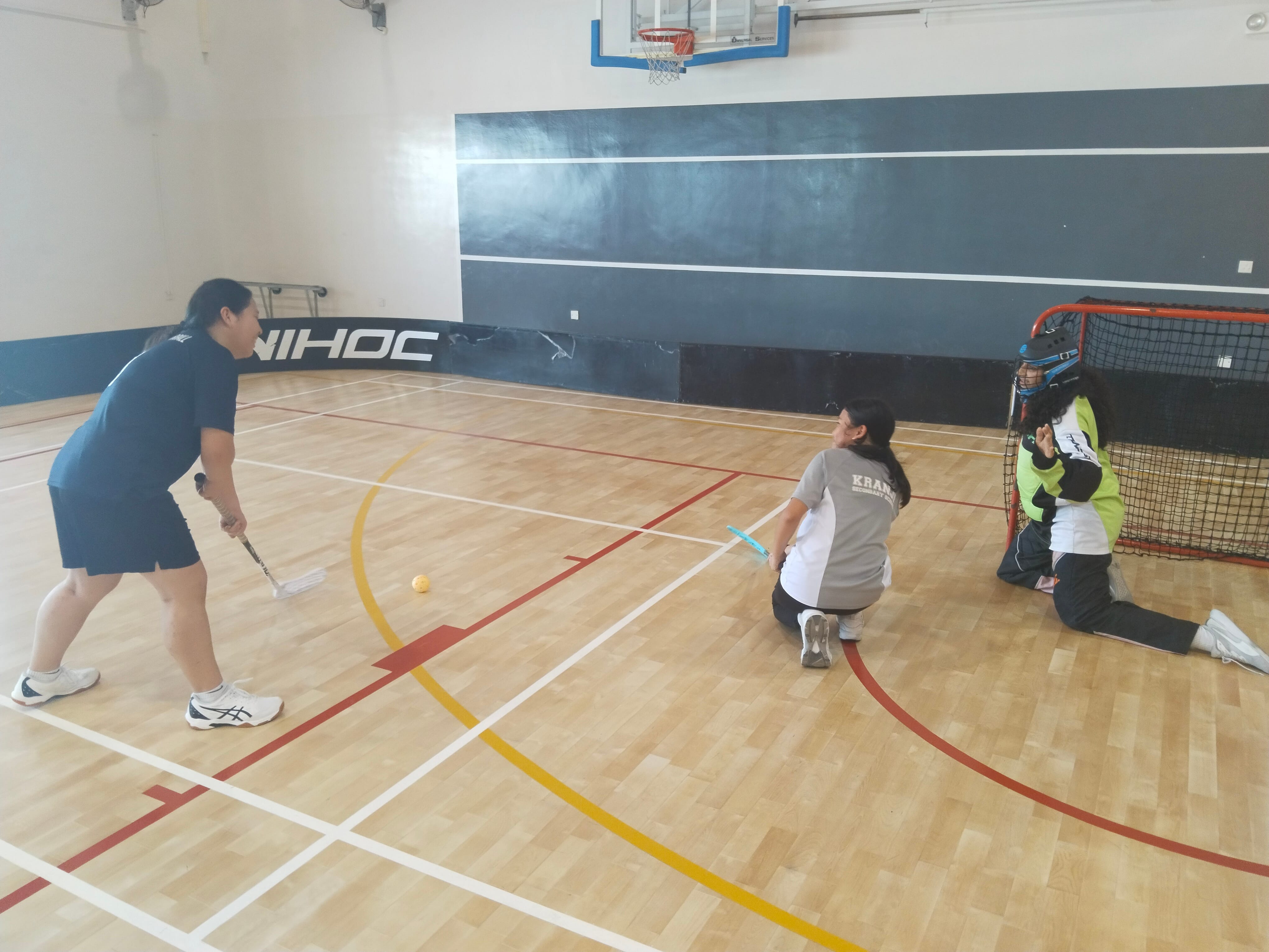 Three people playing floorball indoors, one shooting, one defending, and a helmeted goalie by a red net.