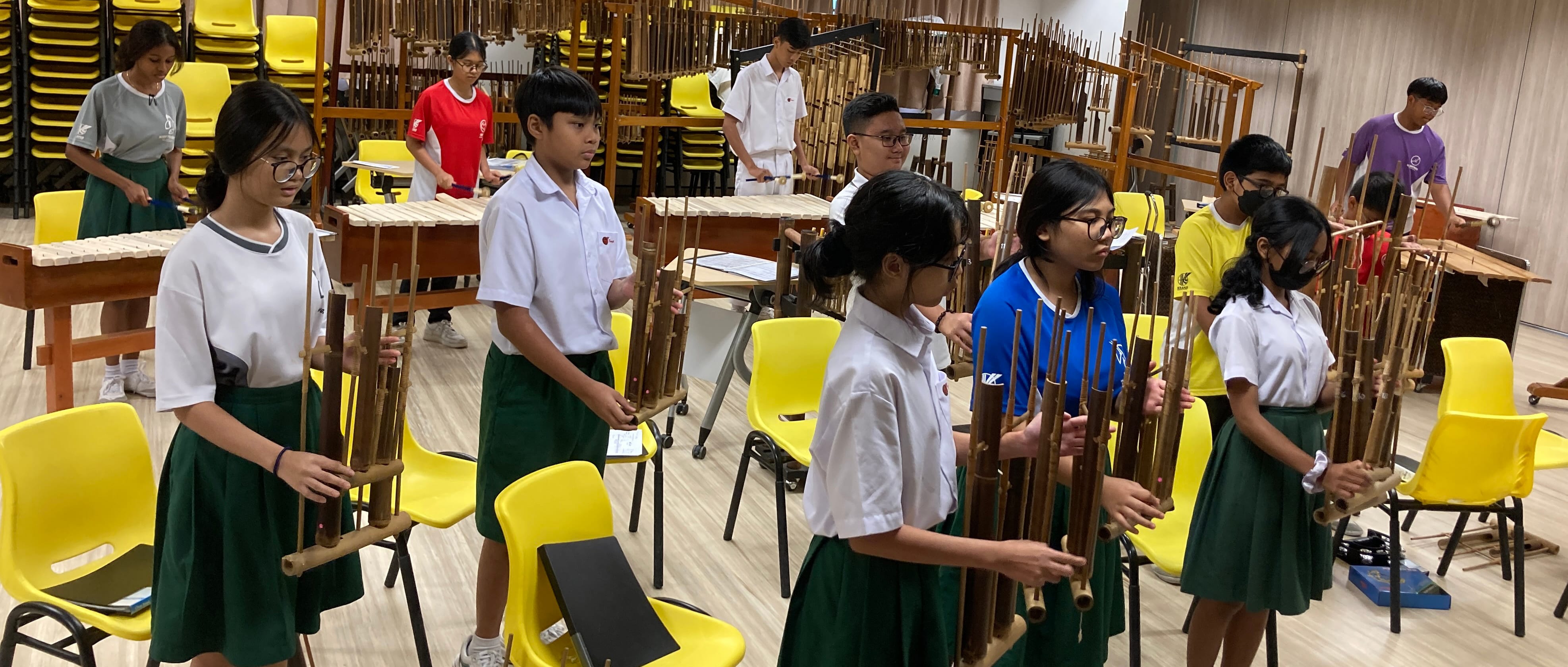 Students in school uniforms play traditional bamboo instruments in a classroom filled with yellow chairs and xylophones.