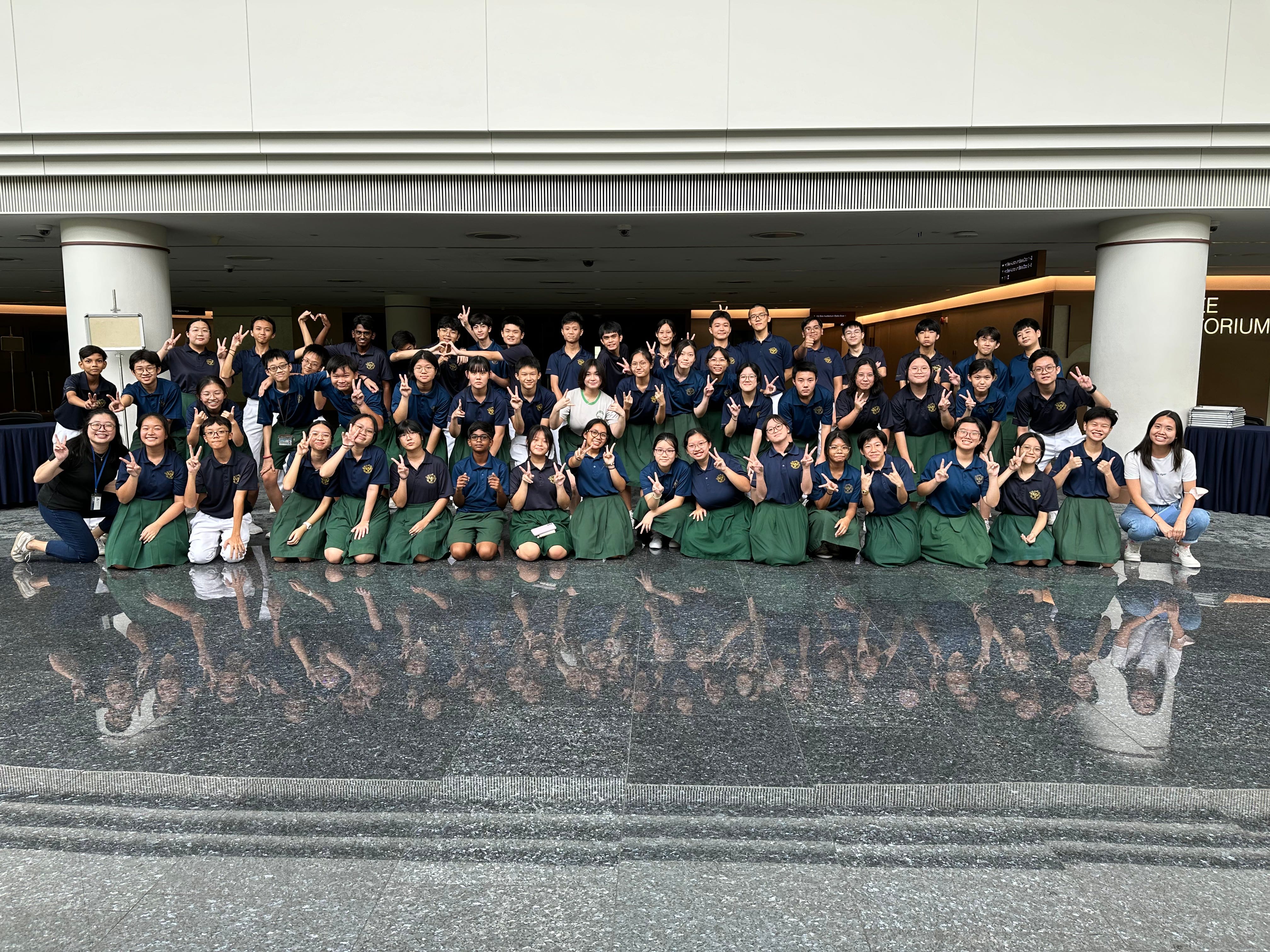 Large group of students in navy uniforms posing with peace signs outside a building with marble floors.