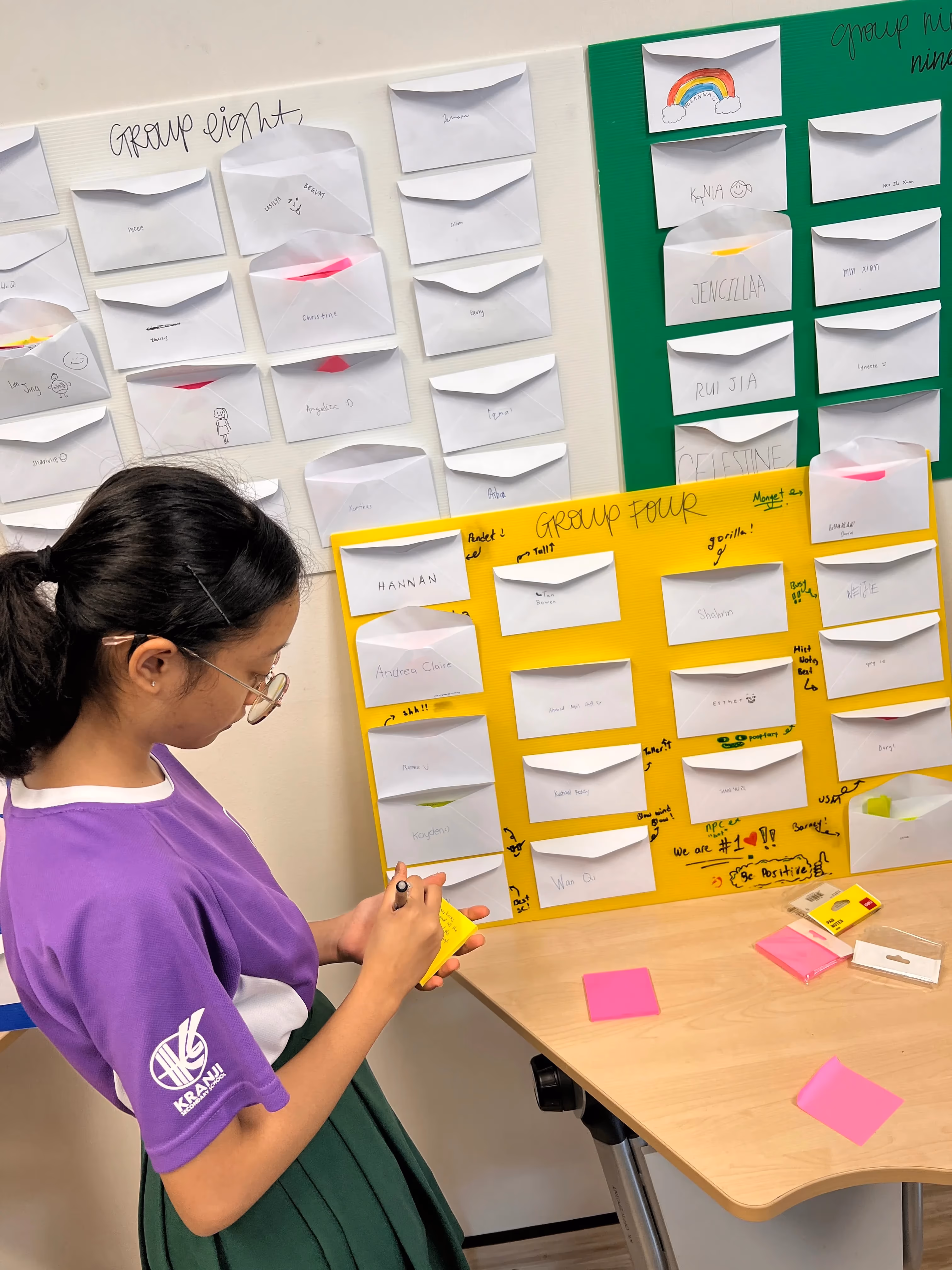 A girl in a purple school uniform writes on a sticky note near boards covered with labeled envelopes for class groups.