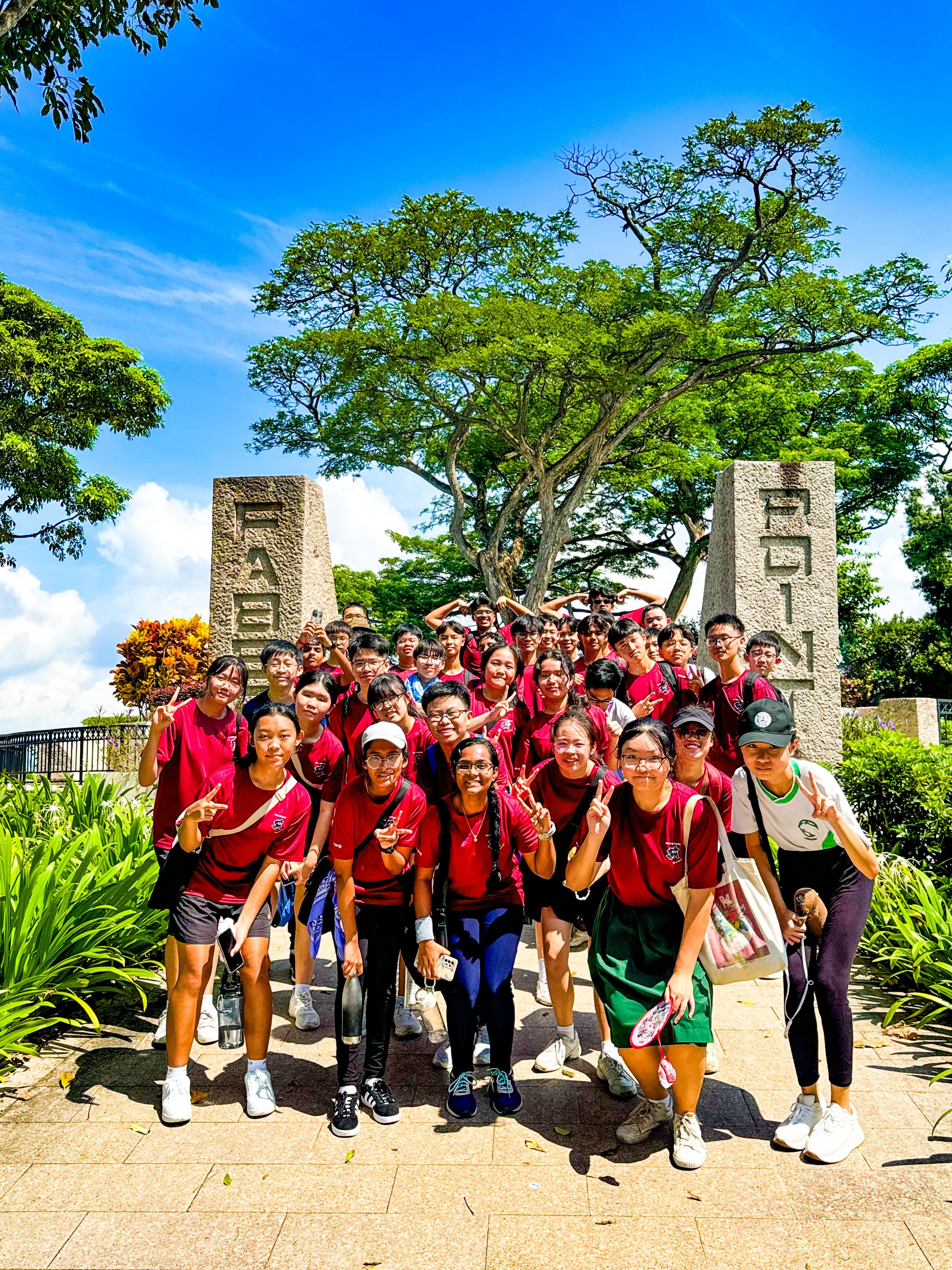 A large group of students in red shirts pose with peace signs between two stone pillars under a large tree.
