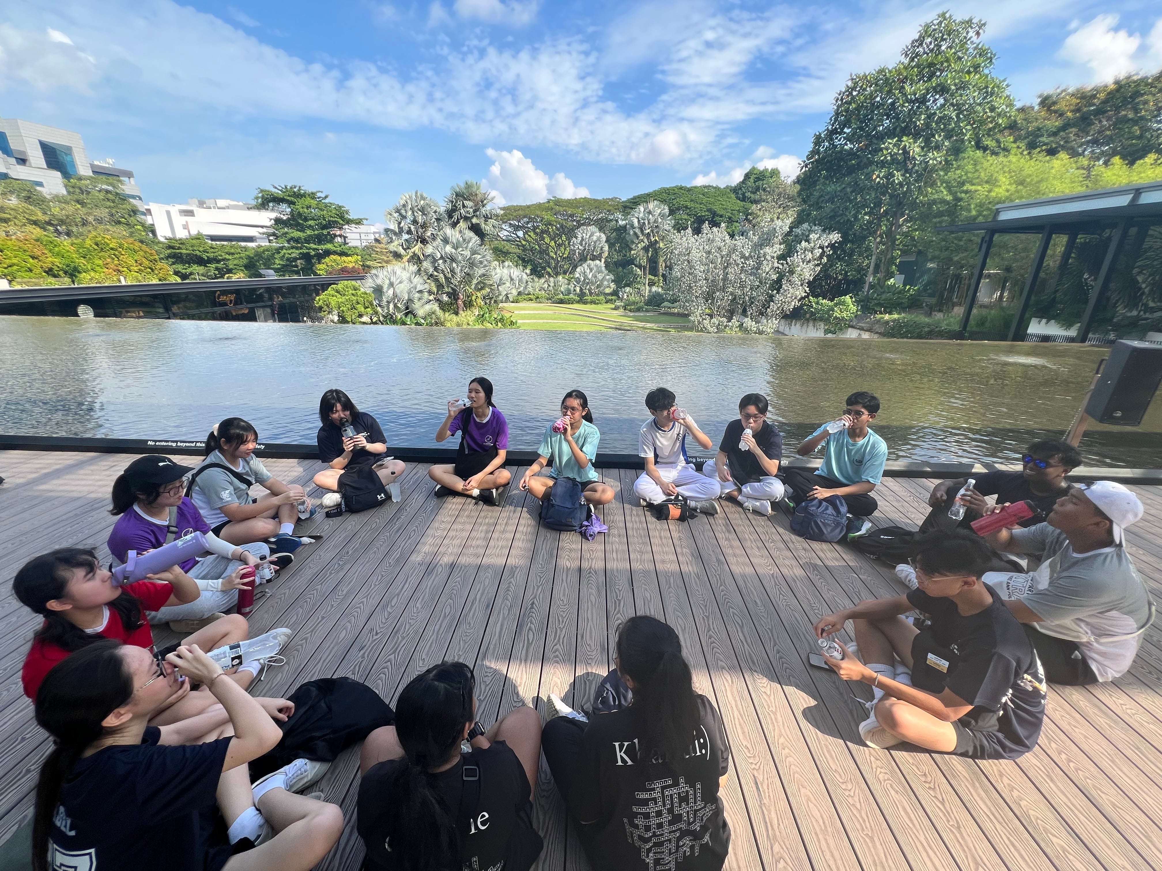 A group of teens sitting in a circle on a wooden deck beside a calm pond surrounded by lush trees.