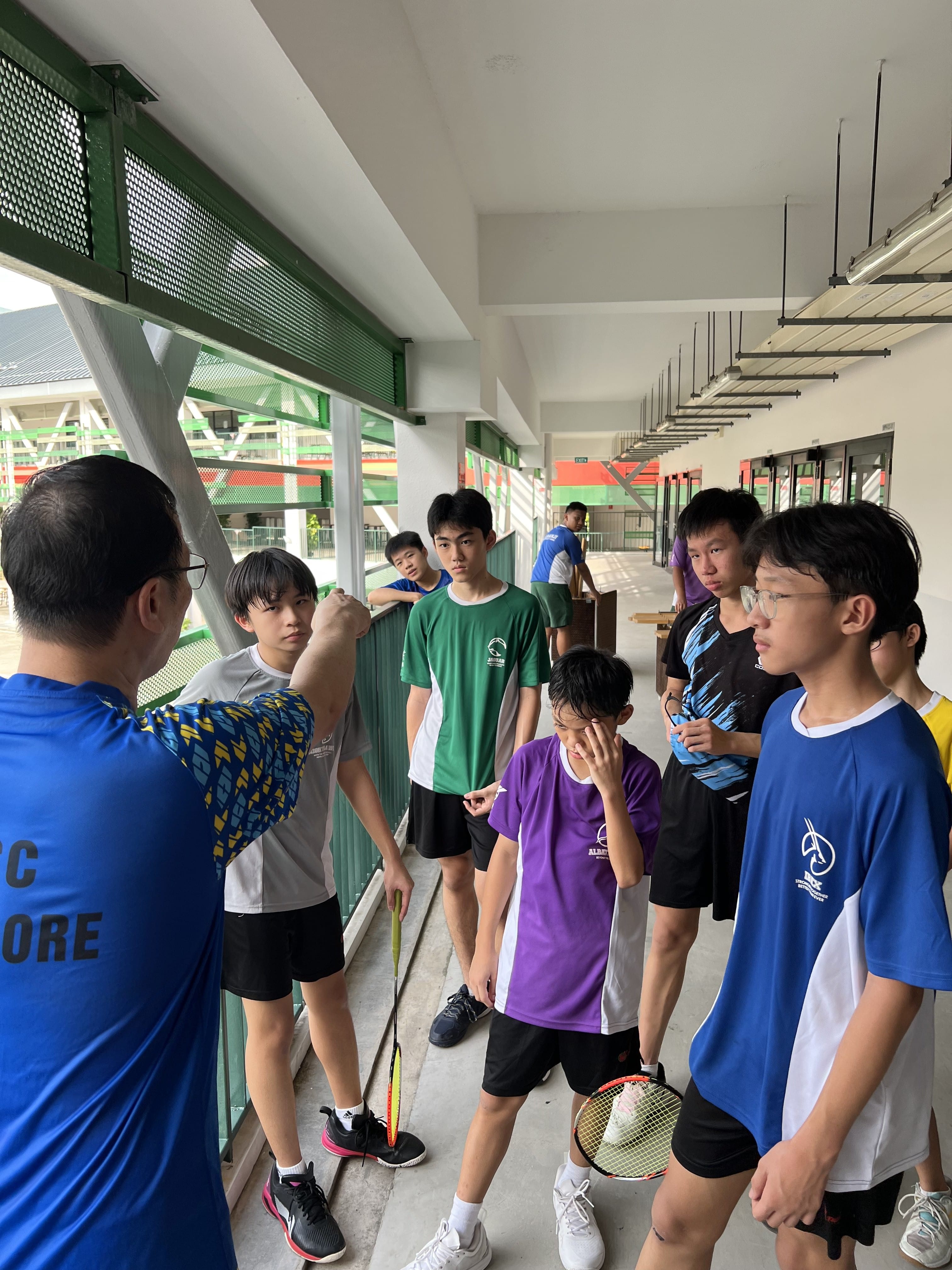 A coach in a blue shirt gestures while instructing a group of young teen badminton players in a covered corridor.