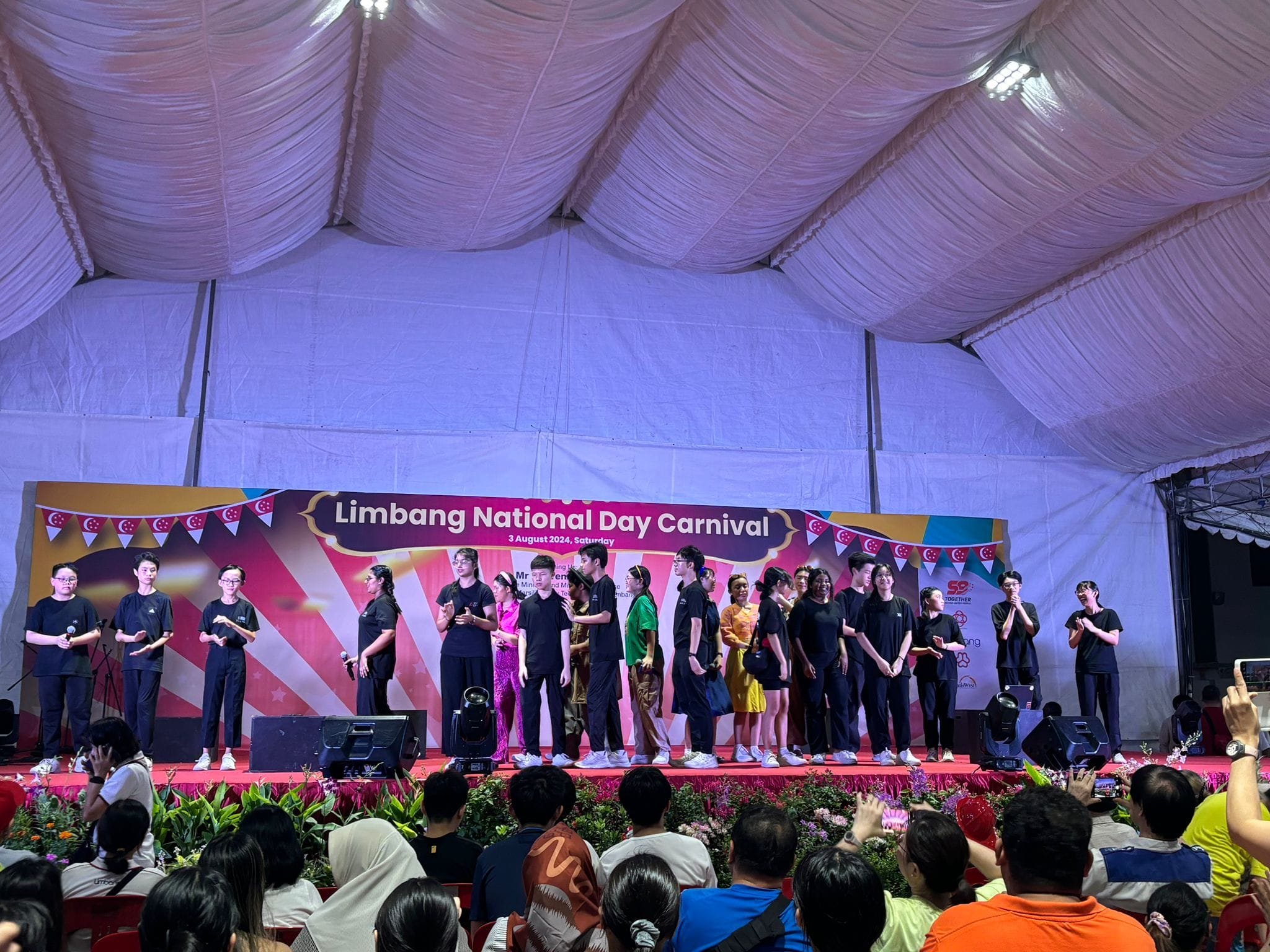 Performers in black stand on a stage at the Limbang National Day Carnival on 3 August 2024, with a crowd watching.