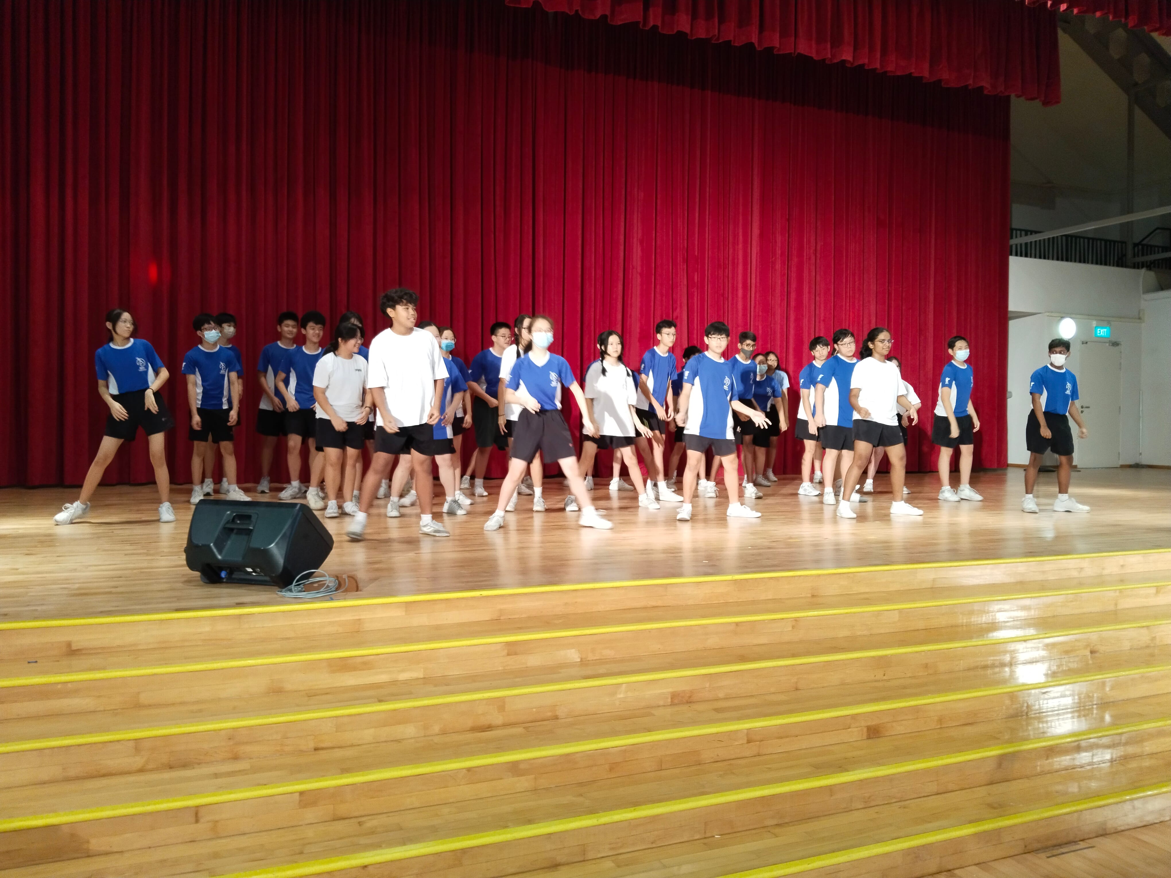 Students in blue and white uniforms performing a dance on a wooden stage in front of a red curtain.