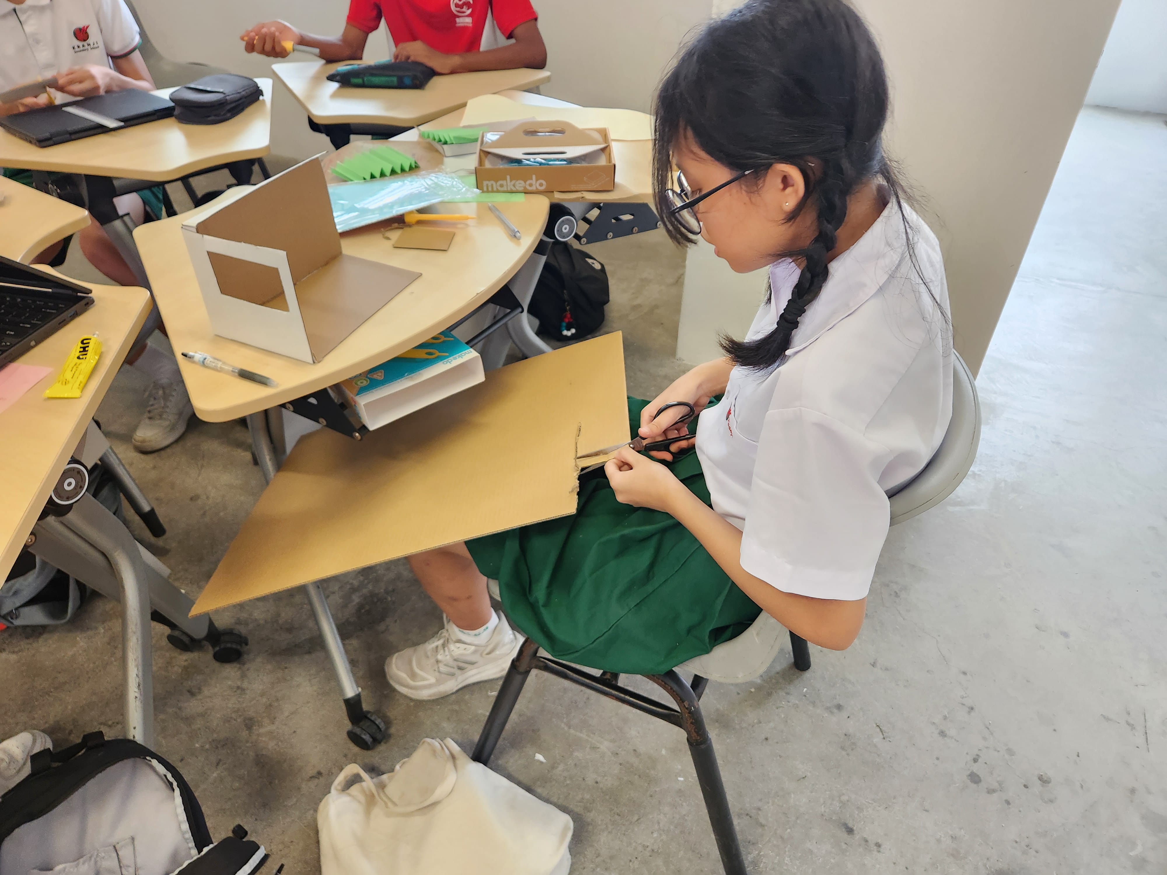 A girl in school uniform cuts cardboard with scissors in a classroom where students build cardboard models.