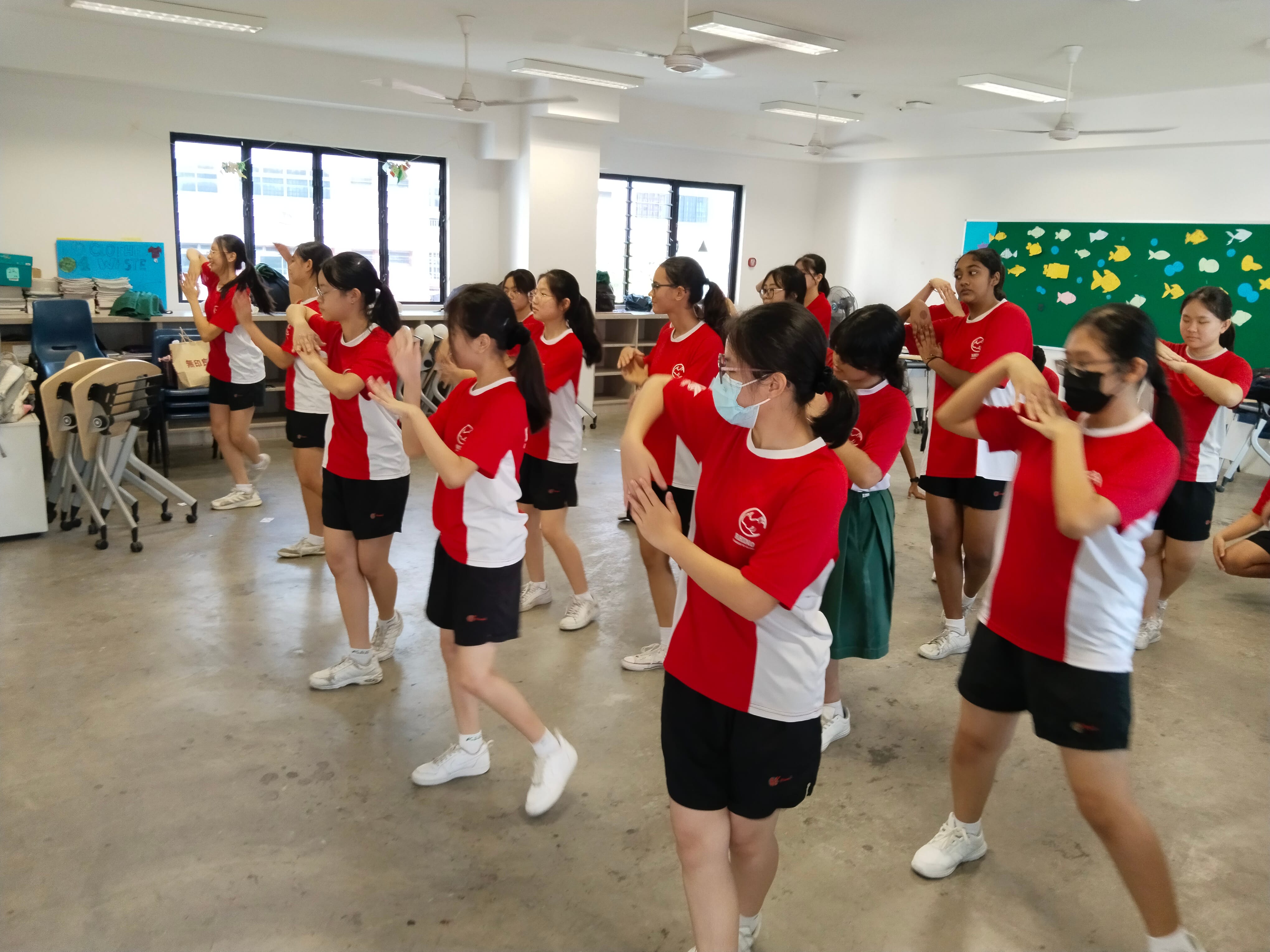 Students in red and white uniforms practice a synchronized dance or exercise routine in a bright indoor classroom.