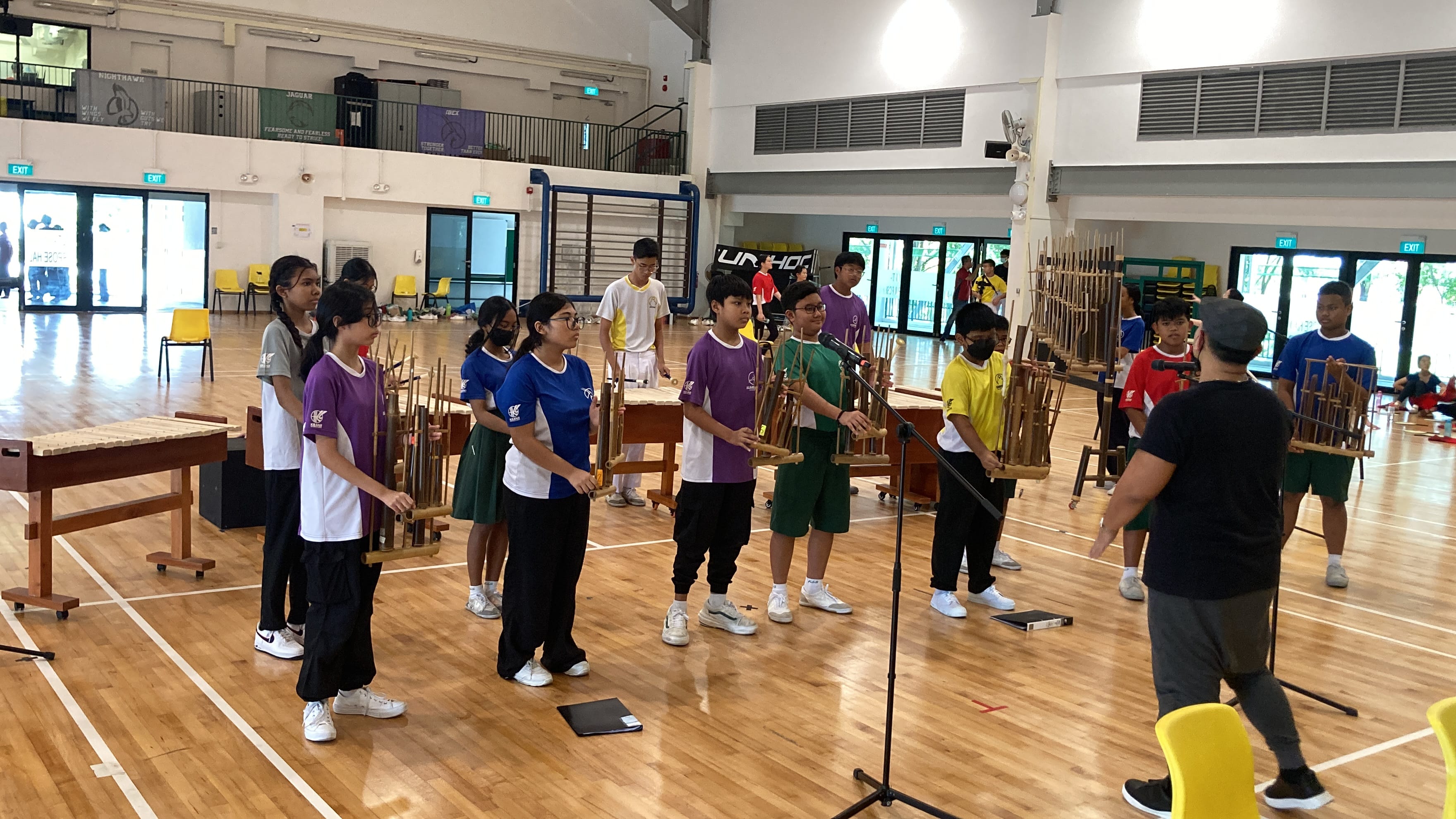 Students in colorful uniforms play traditional bamboo instruments in a school gymnasium led by an instructor at a microphone.