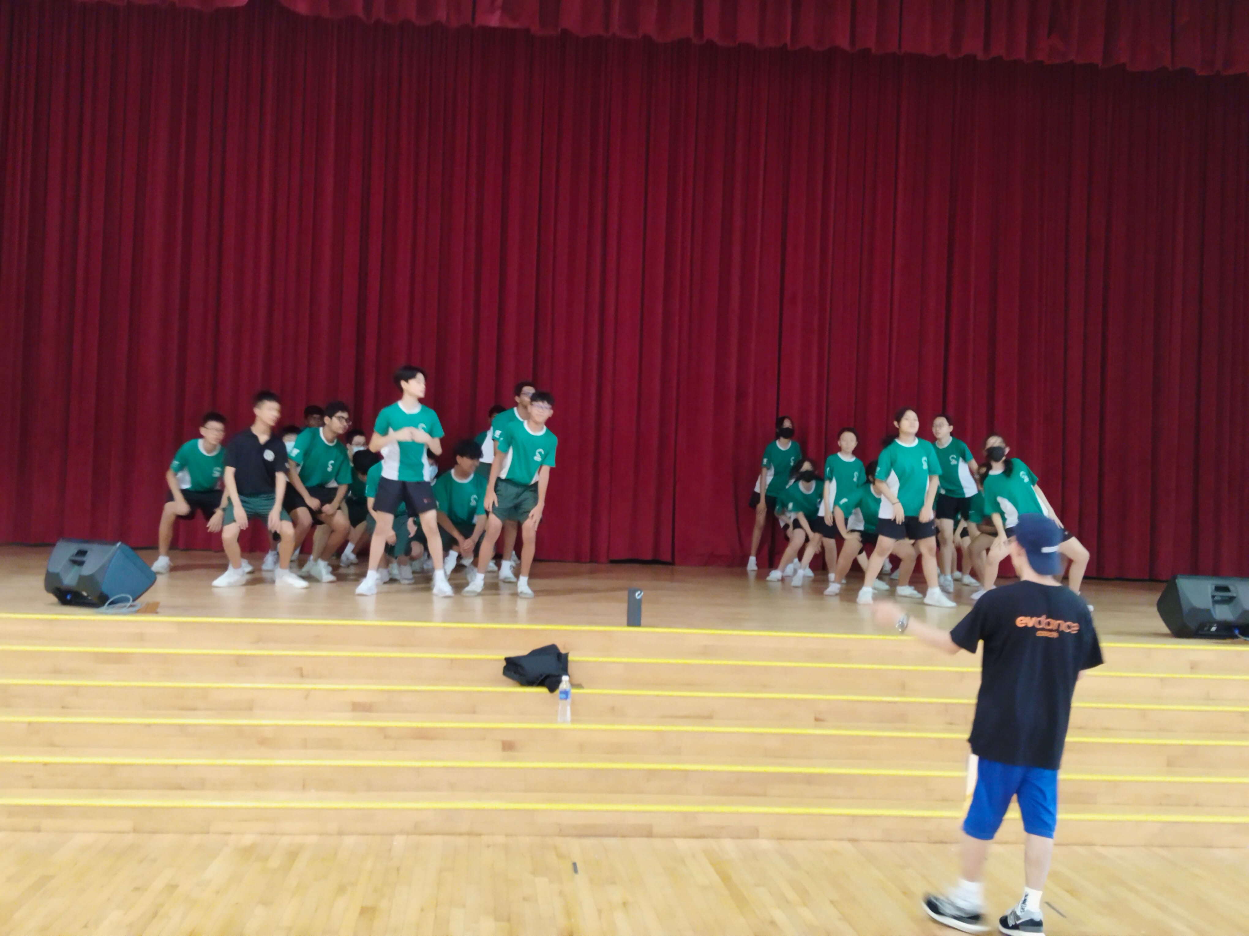 A dance instructor in black directs a large group of students in green shirts rehearsing on a stage with red curtains.