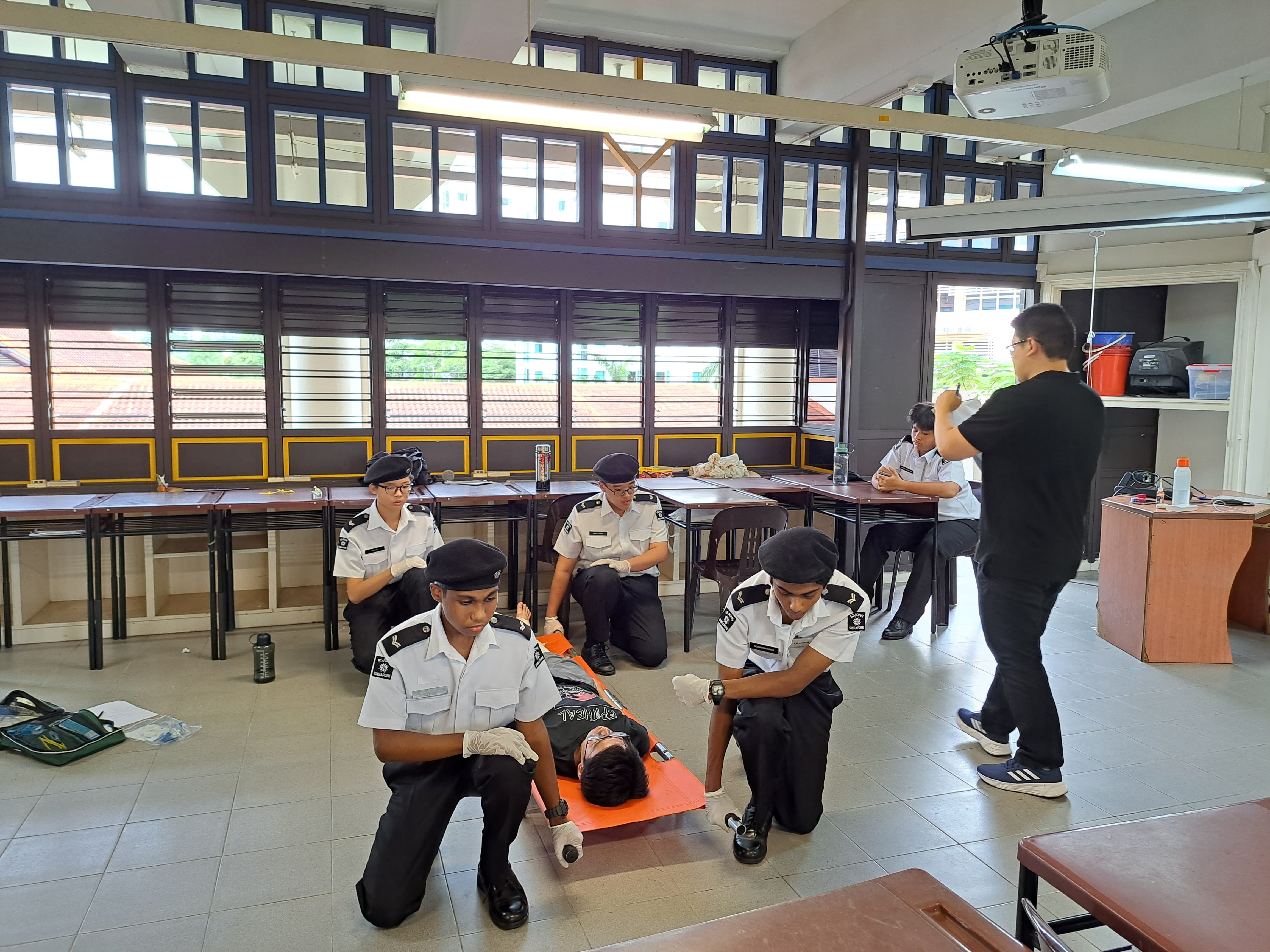 Uniformed cadets kneel around a person on an orange stretcher as an instructor stands nearby in a classroom.