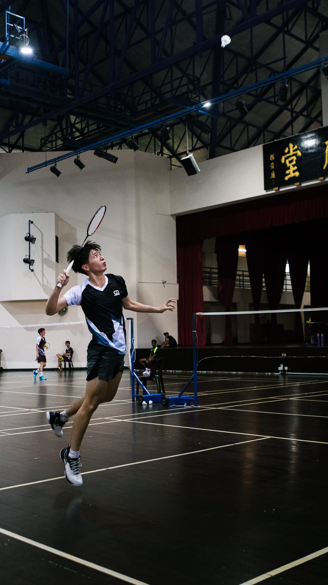 Young male player jumps mid-air swinging a badminton racket toward a shuttlecock inside an indoor court.
