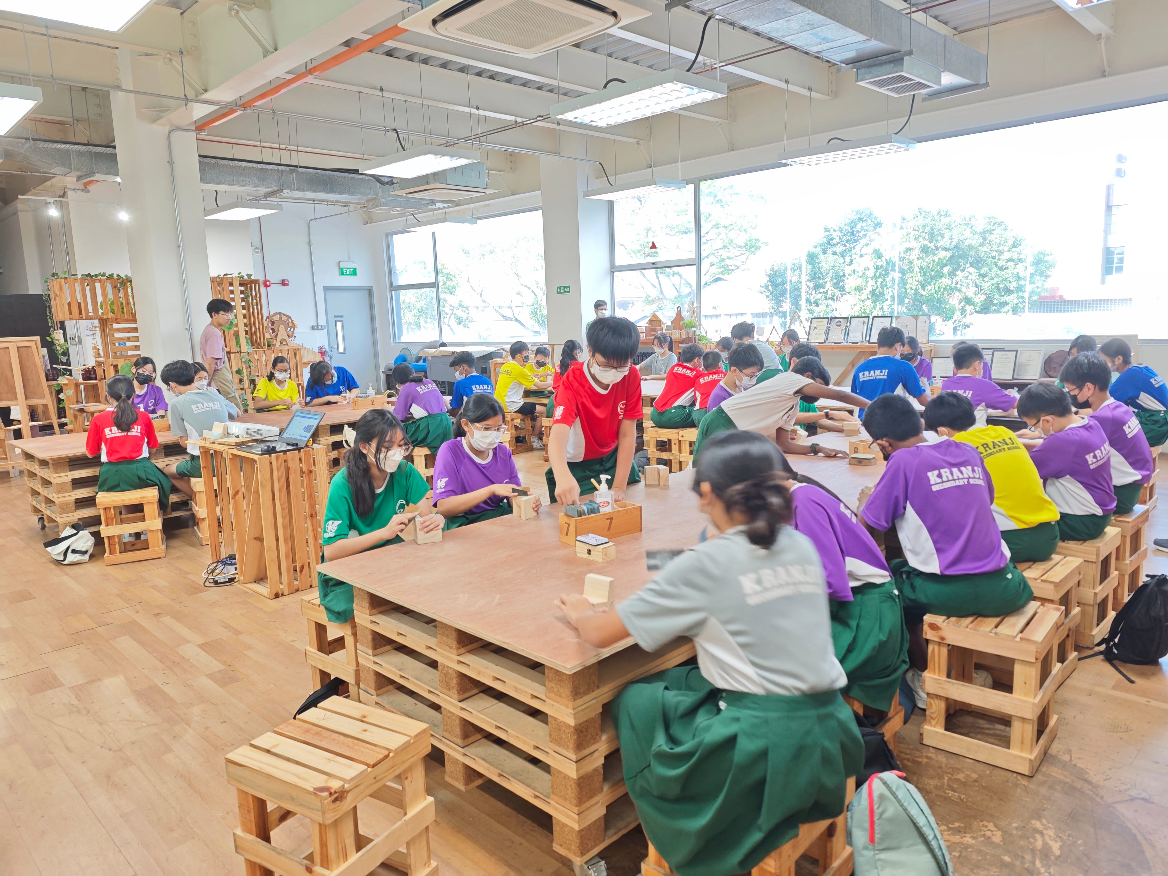 Students in colorful uniforms sit at wooden pallet tables working with small wooden blocks in a bright workshop room.