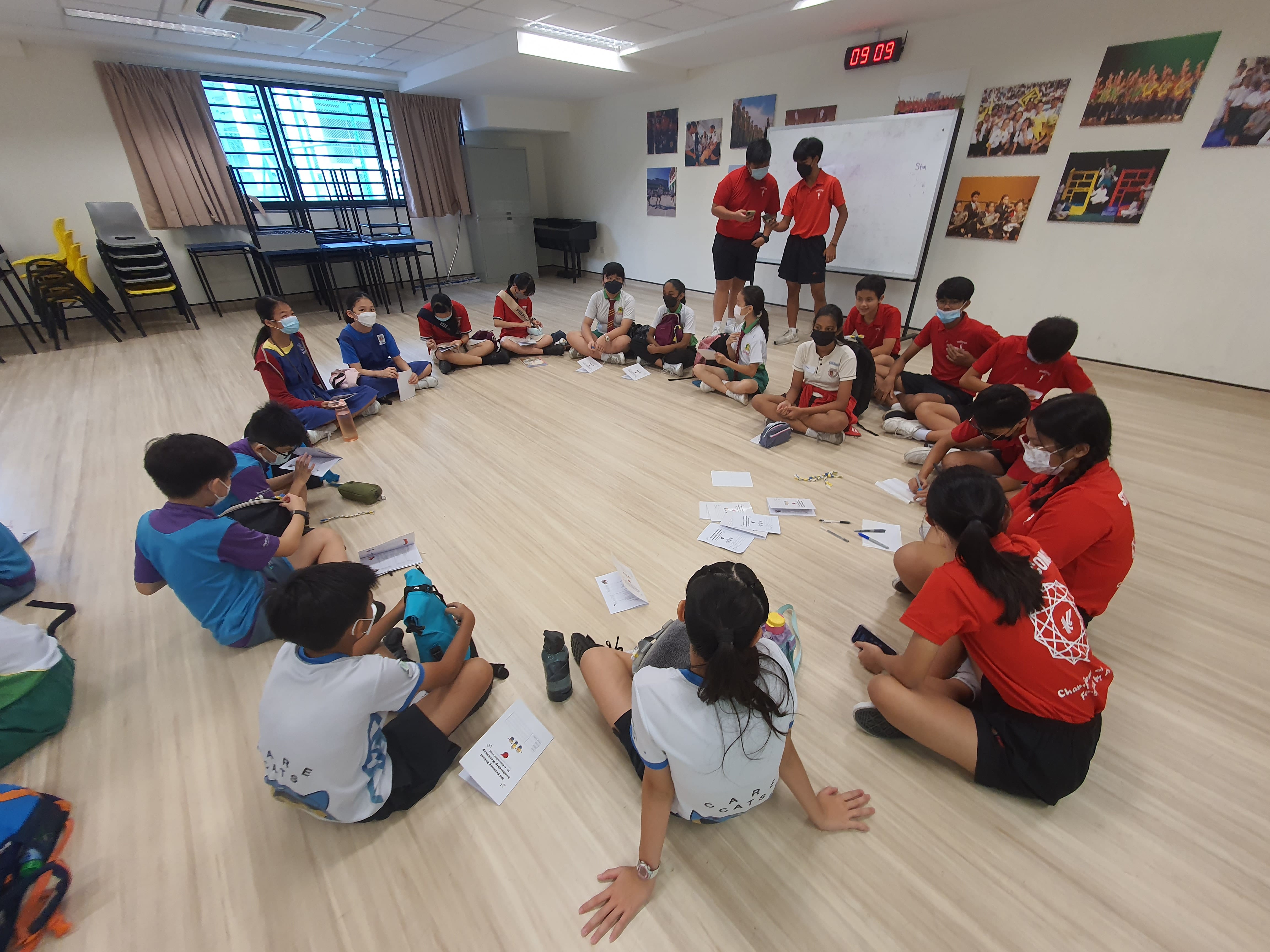 Students wearing masks sit in a circle on a wooden floor in a classroom, with papers and two standing leaders nearby.