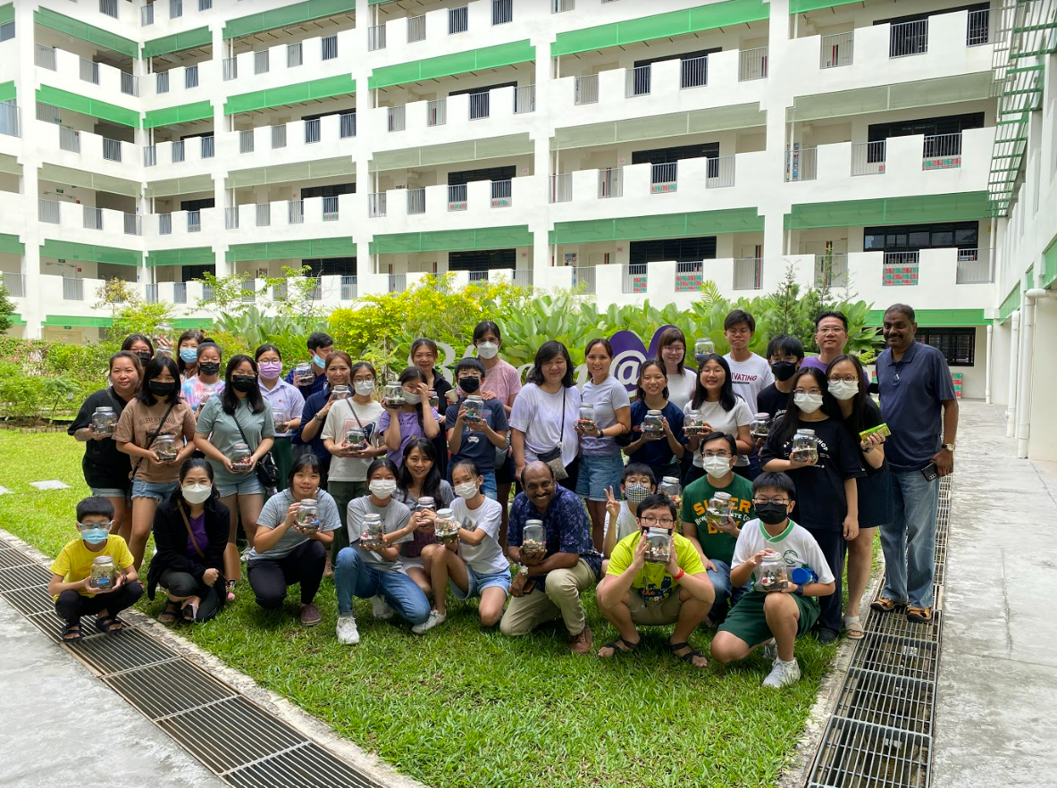 A large group of masked people outdoors holding glass jars, posing in front of a white and green multi-story building.