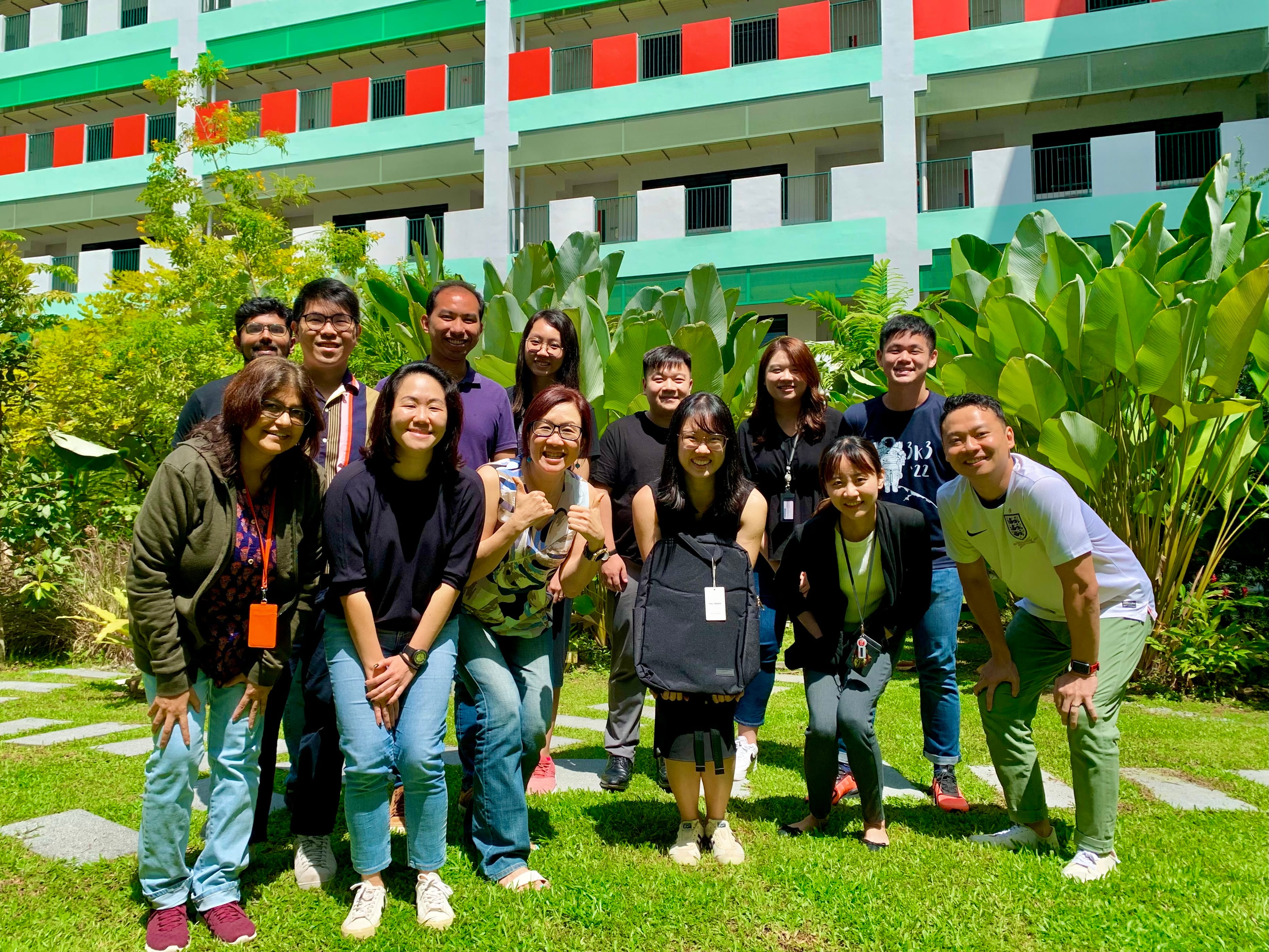 A group of about 13 people smiling outdoors on grass in front of a green and red multi-story building.