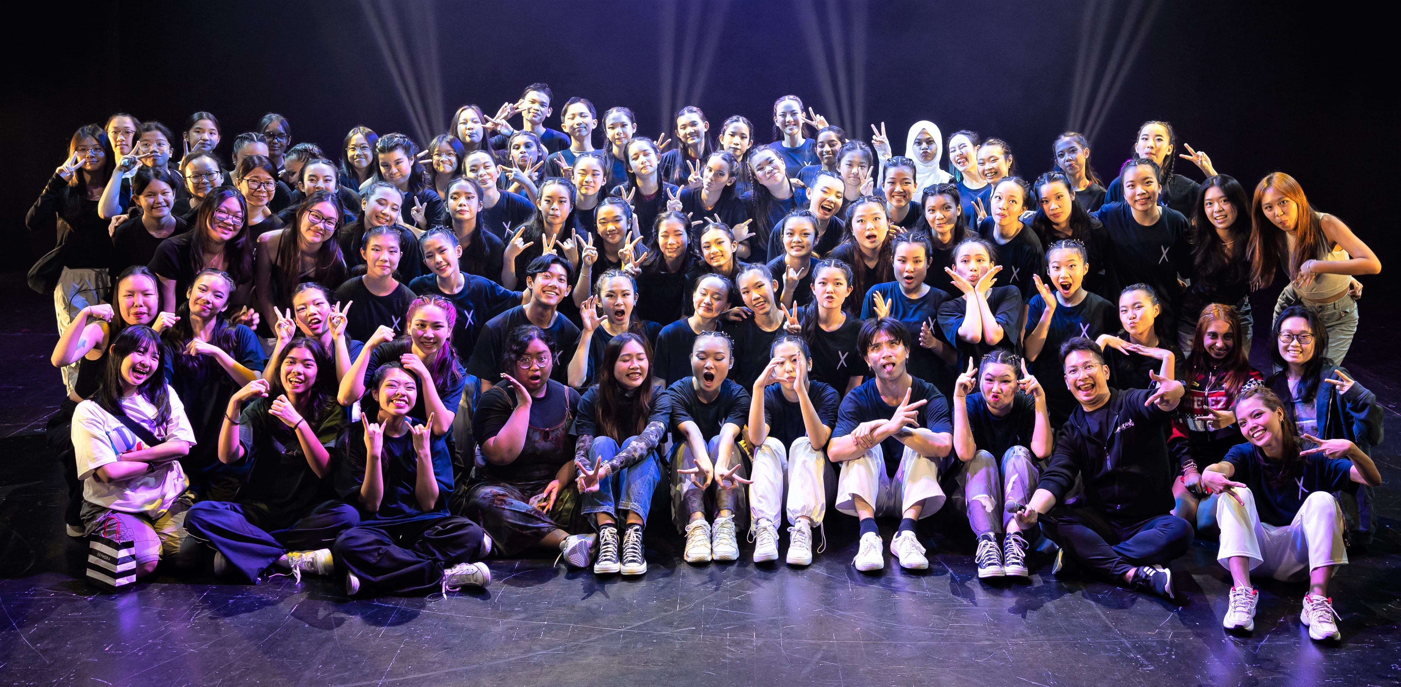 Large group of young people mostly in black shirts posing on a stage with bright spotlights, making hand signs.