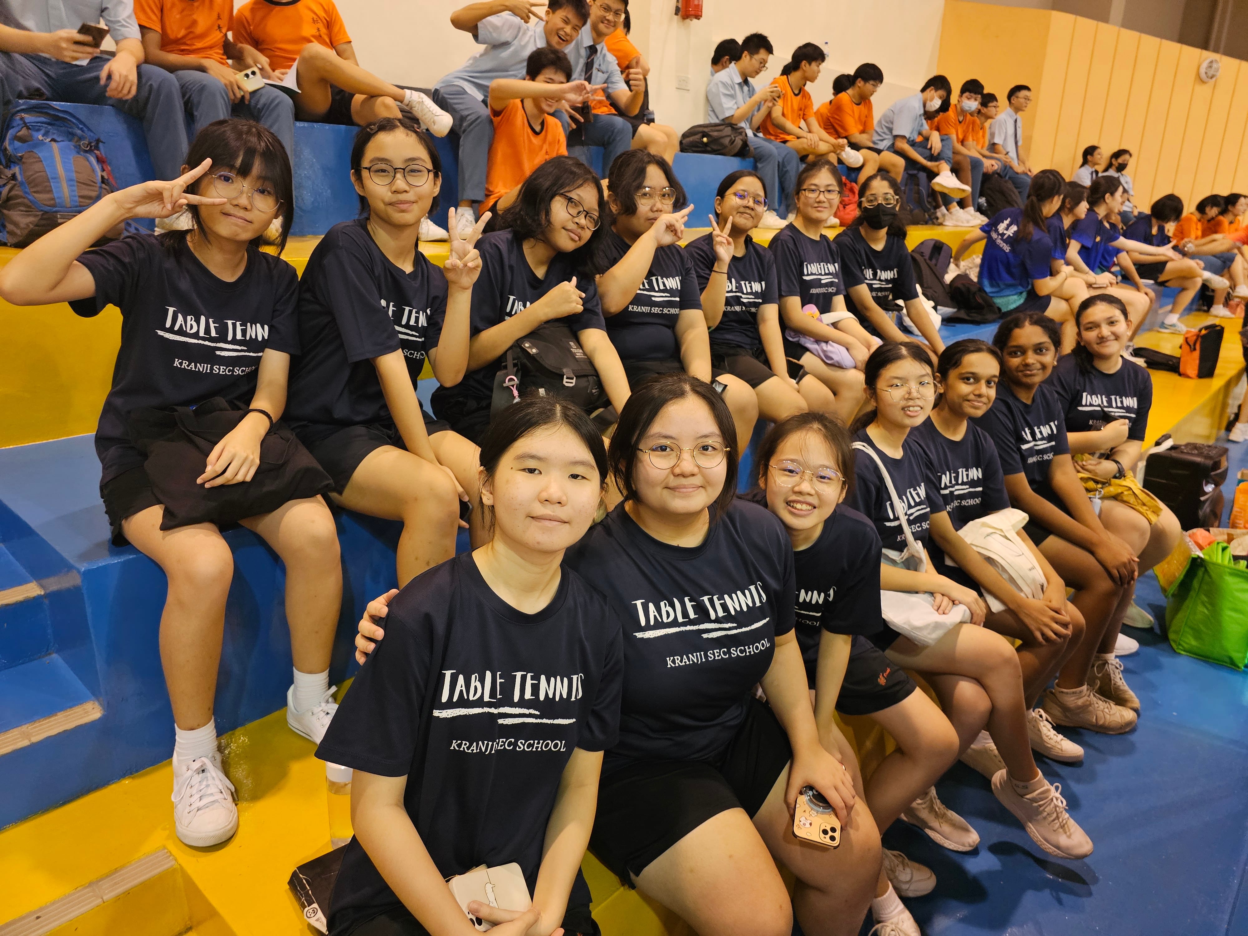 A group of girls in navy Table Tennis Kranji Sec School shirts sit on bleachers, smiling and making peace signs.