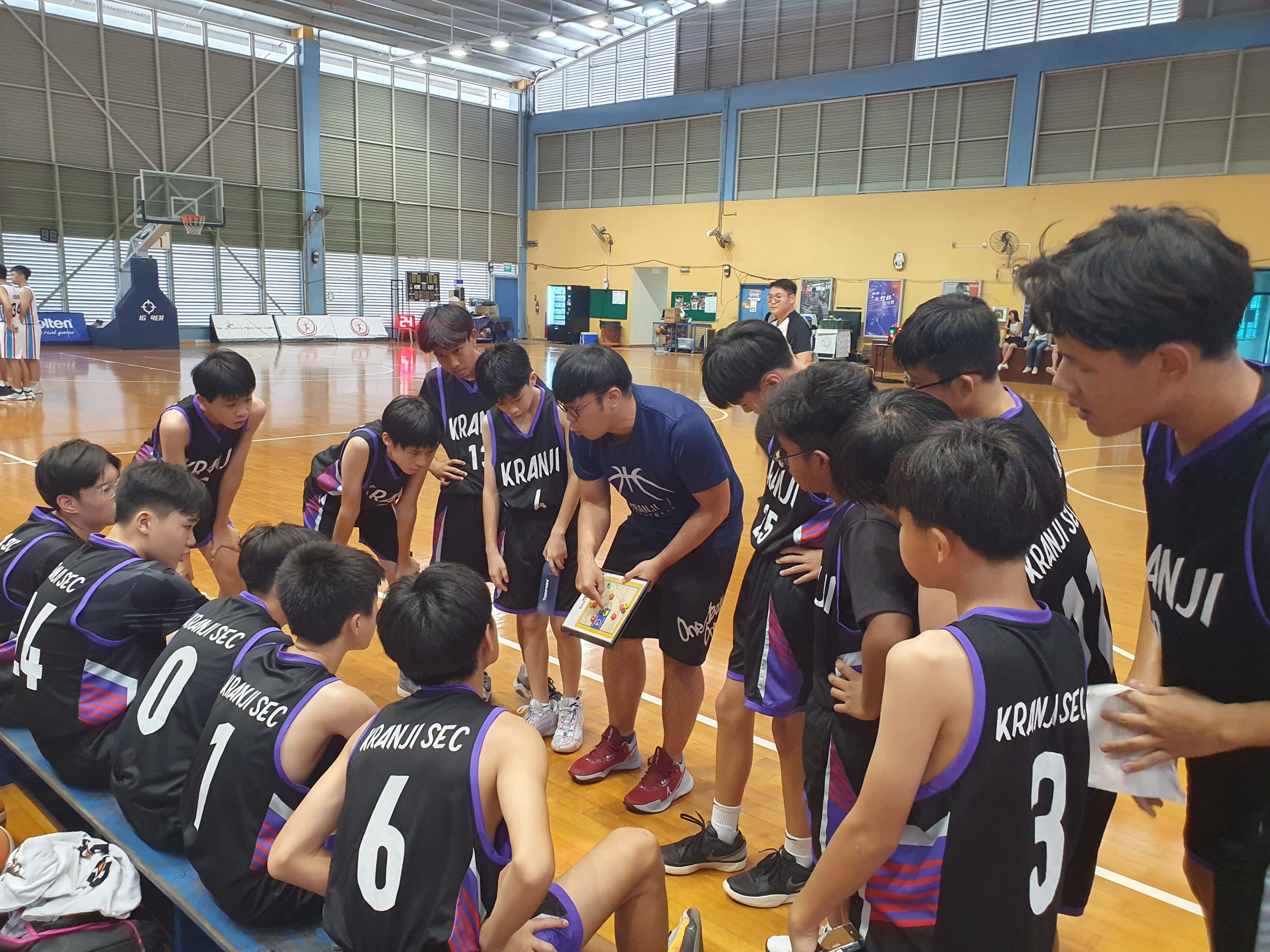 A coach holds a clipboard and talks to Kranji Secondary School basketball players huddled together in an indoor court.