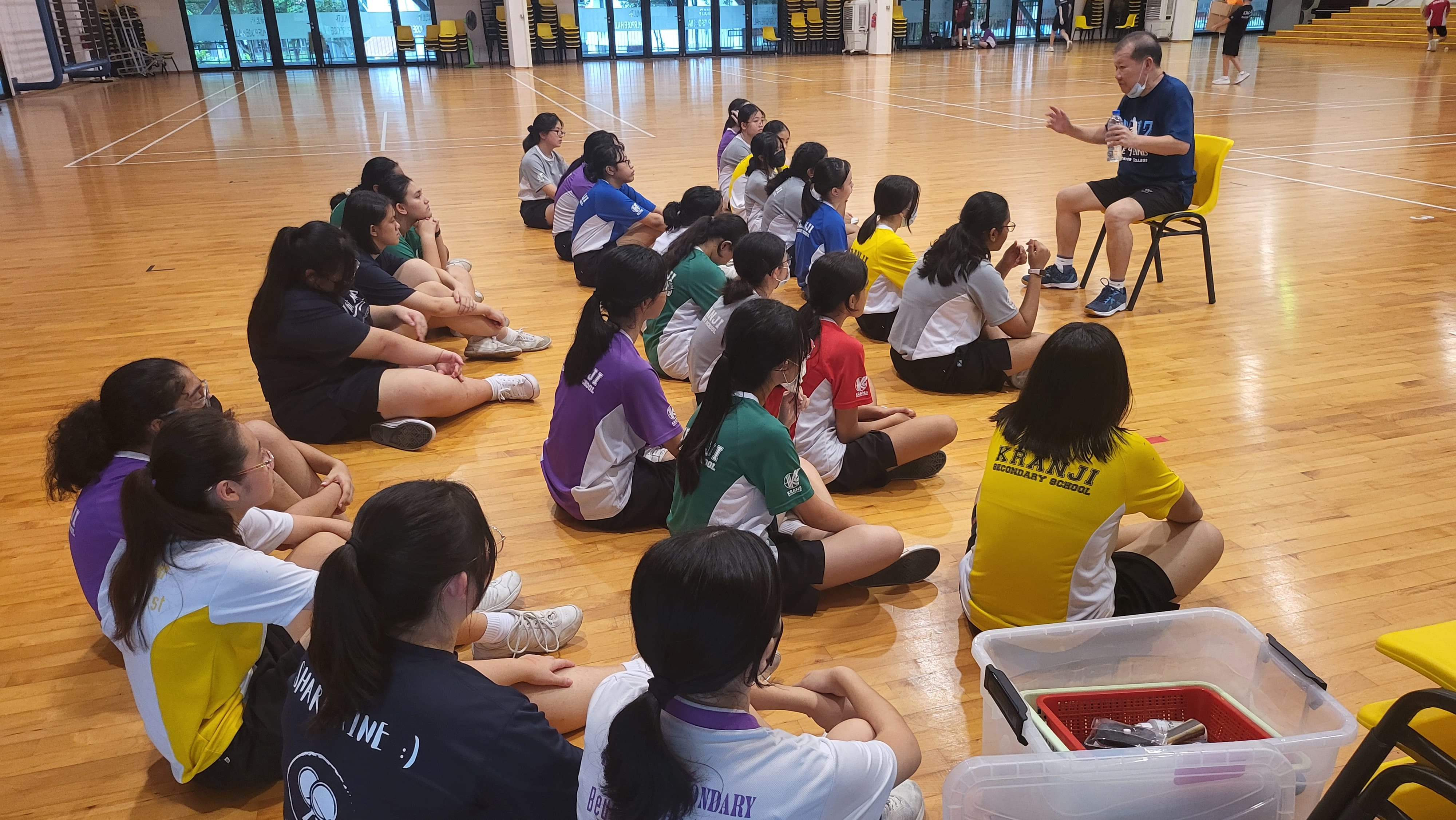 A coach sits on a chair addressing a group of students seated on a wooden gym floor in colorful school jerseys.