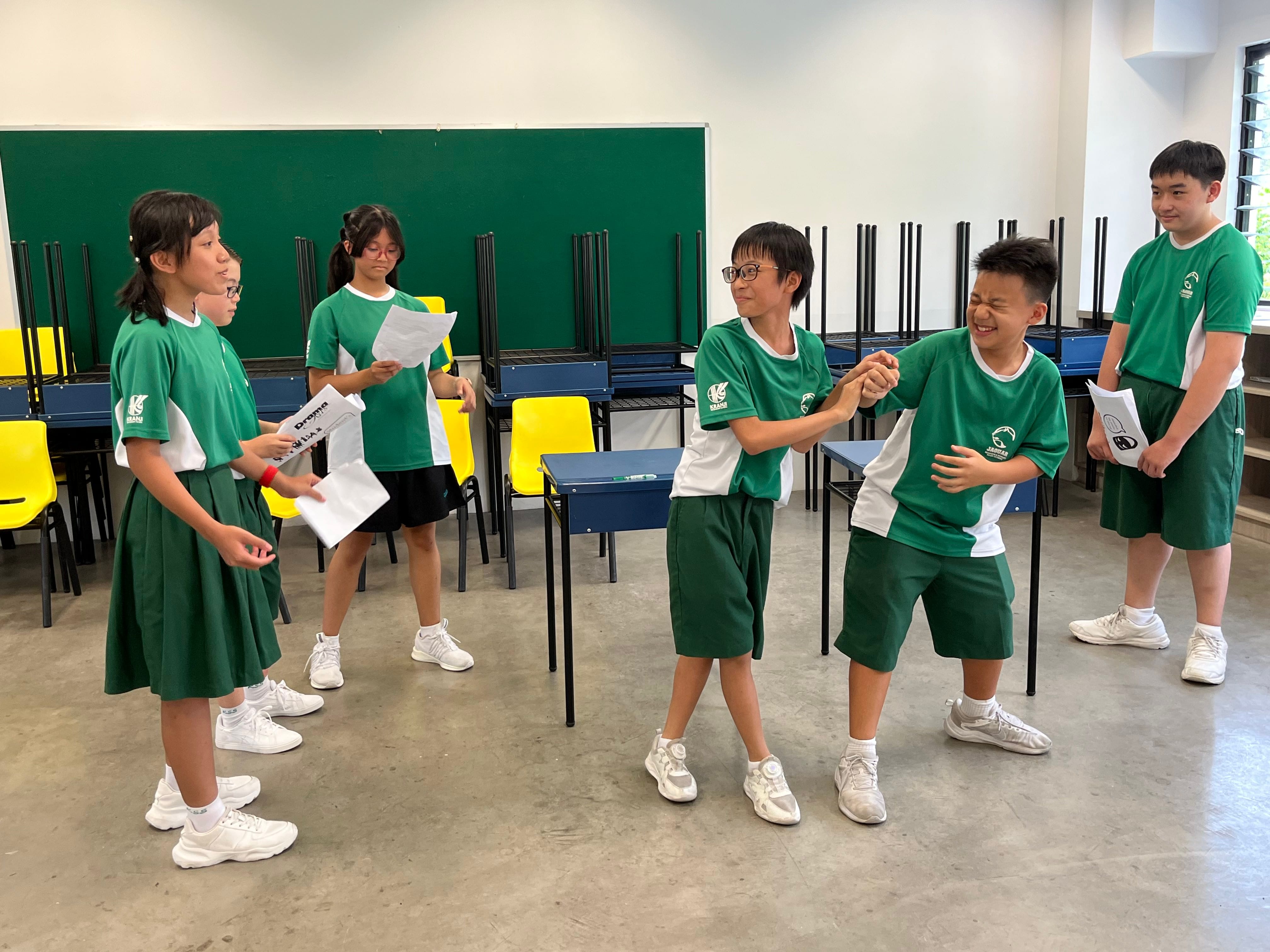 Five students in green school uniforms rehearse a drama scene in a classroom, holding scripts and acting out roles.