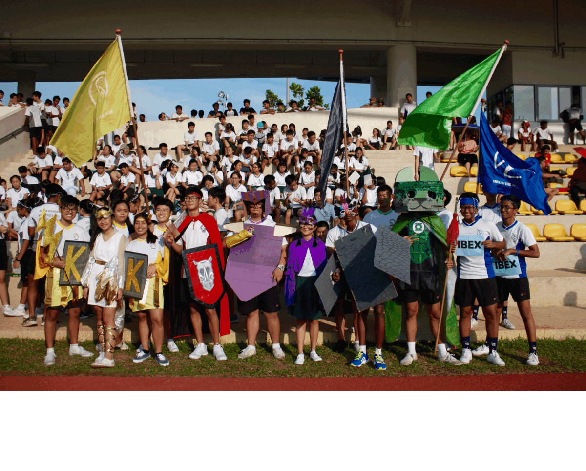 Students in costume hold colorful flags on a track, with packed bleachers behind them.