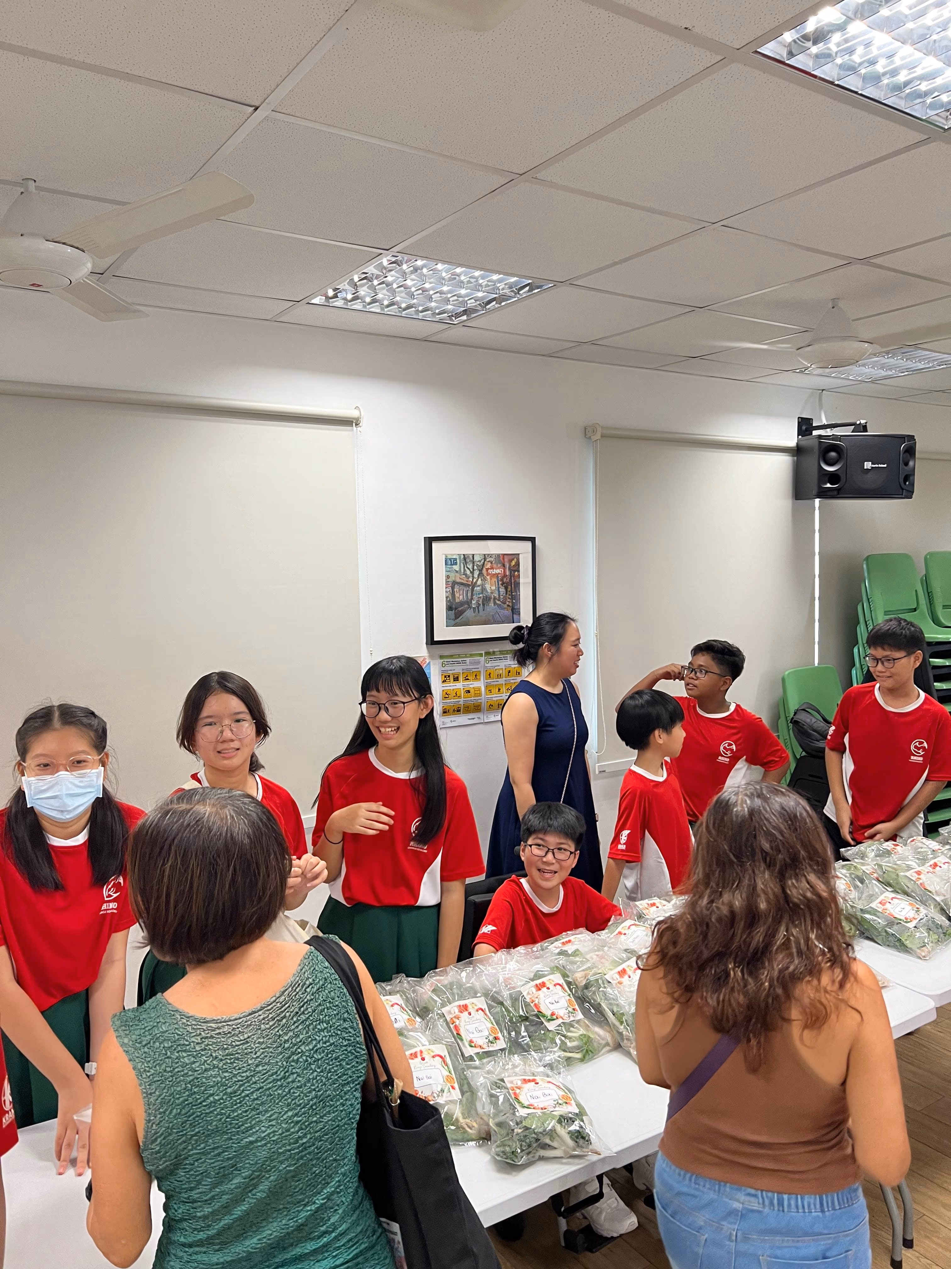 Students in red uniforms stand around a table with bagged vegetables, interacting with adults in an indoor room.