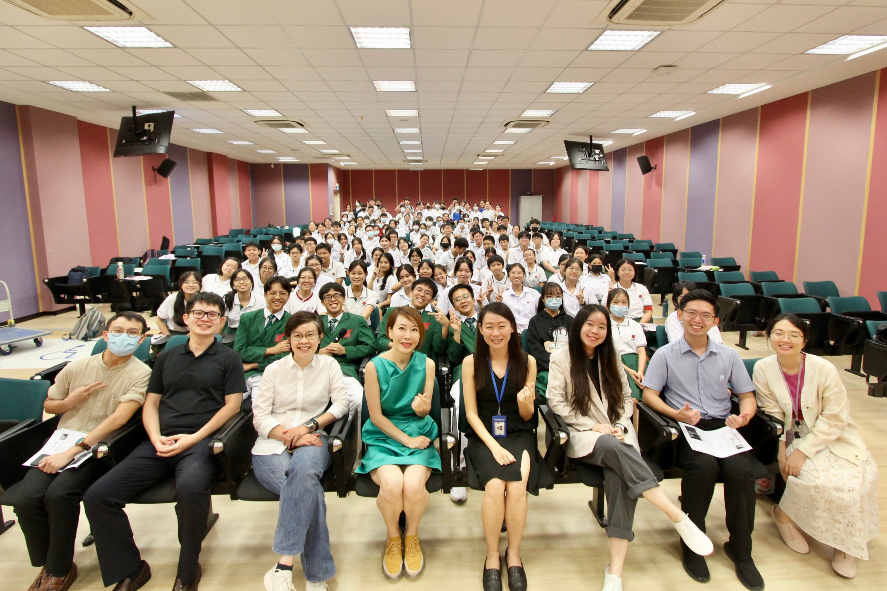 Large group of students in white uniforms and adults posing together in a tiered lecture hall with pink and purple walls.